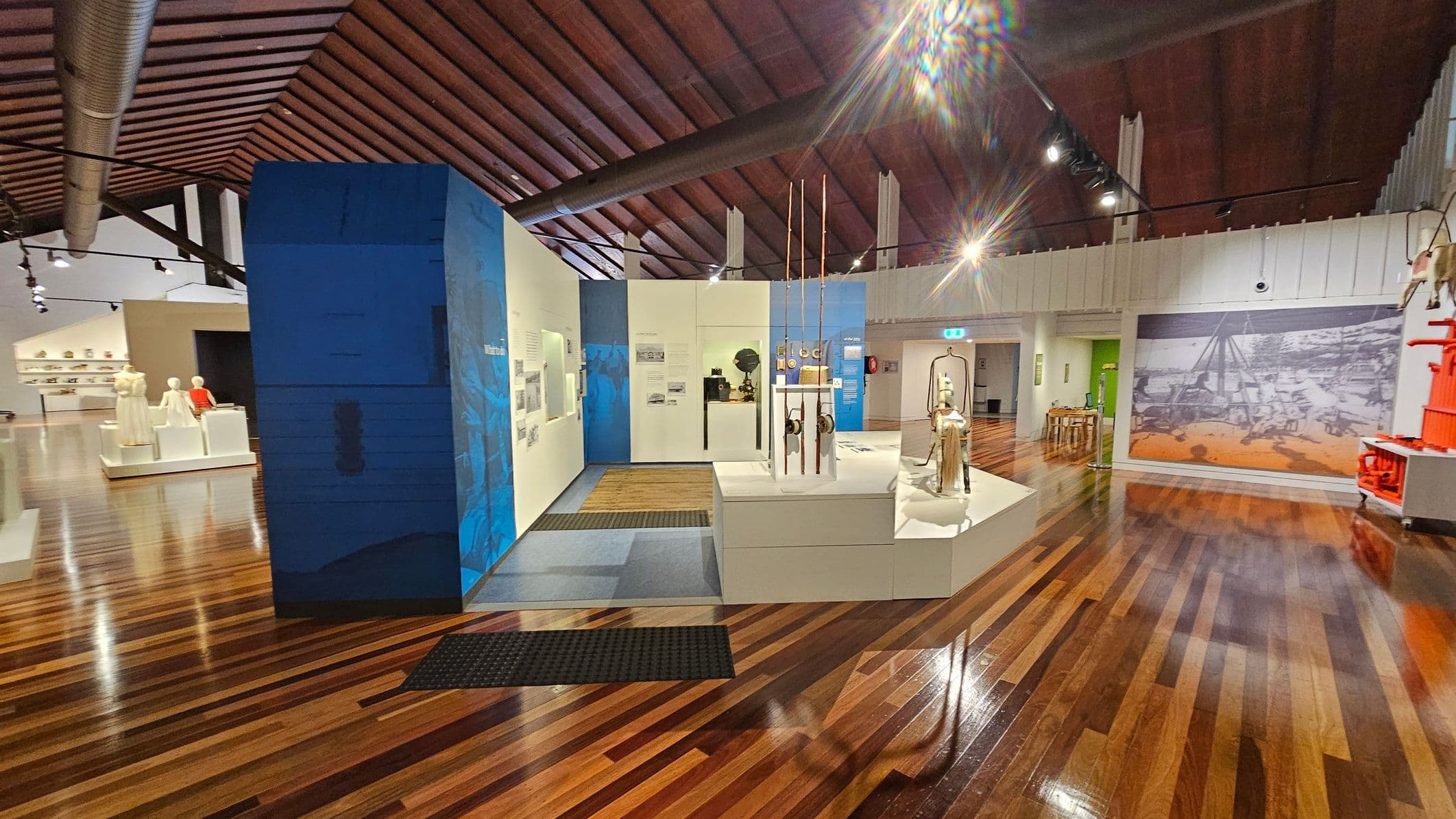 A museum interior with polished wooden floors and a high, slatted wooden ceiling. In the centre, there are display cases with various artefacts, including tall, thin objects and a mannequin. The left side features a blue wall with text and images, while the right side has a large mural and a red structure. In the background, there are more displays and a small seating area. Overhead lighting illuminates the exhibits.