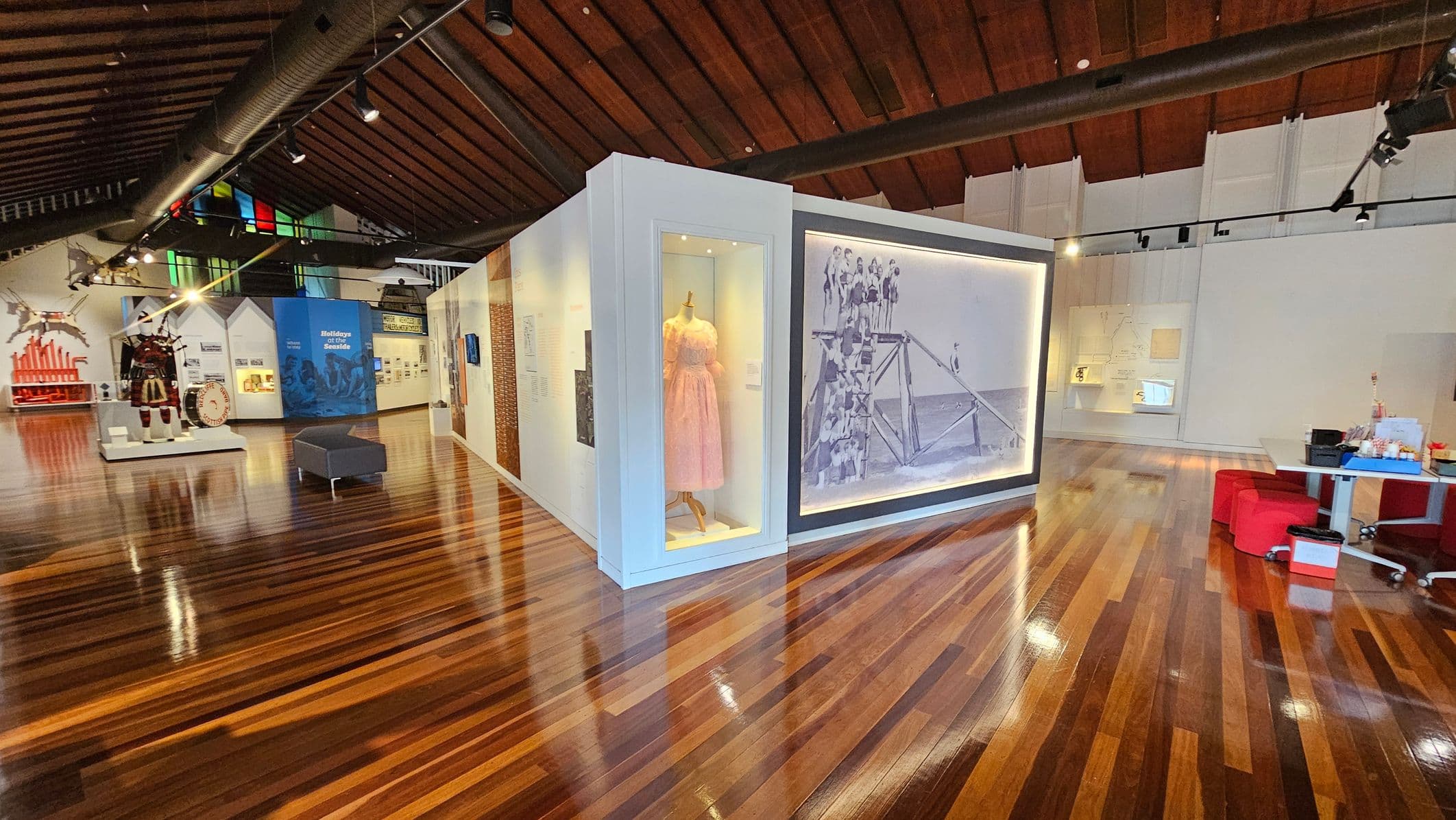 A museum interior with polished wooden floors and a high, wooden ceiling. On the left, there are displays including a mannequin in a traditional outfit and a large red organ. In the centre, a glass case contains a pink dress, and a large black-and-white photo is mounted on the wall. On the right, there are red seating areas and a small table with items on it. Stained glass windows and various exhibits are visible in the background.
