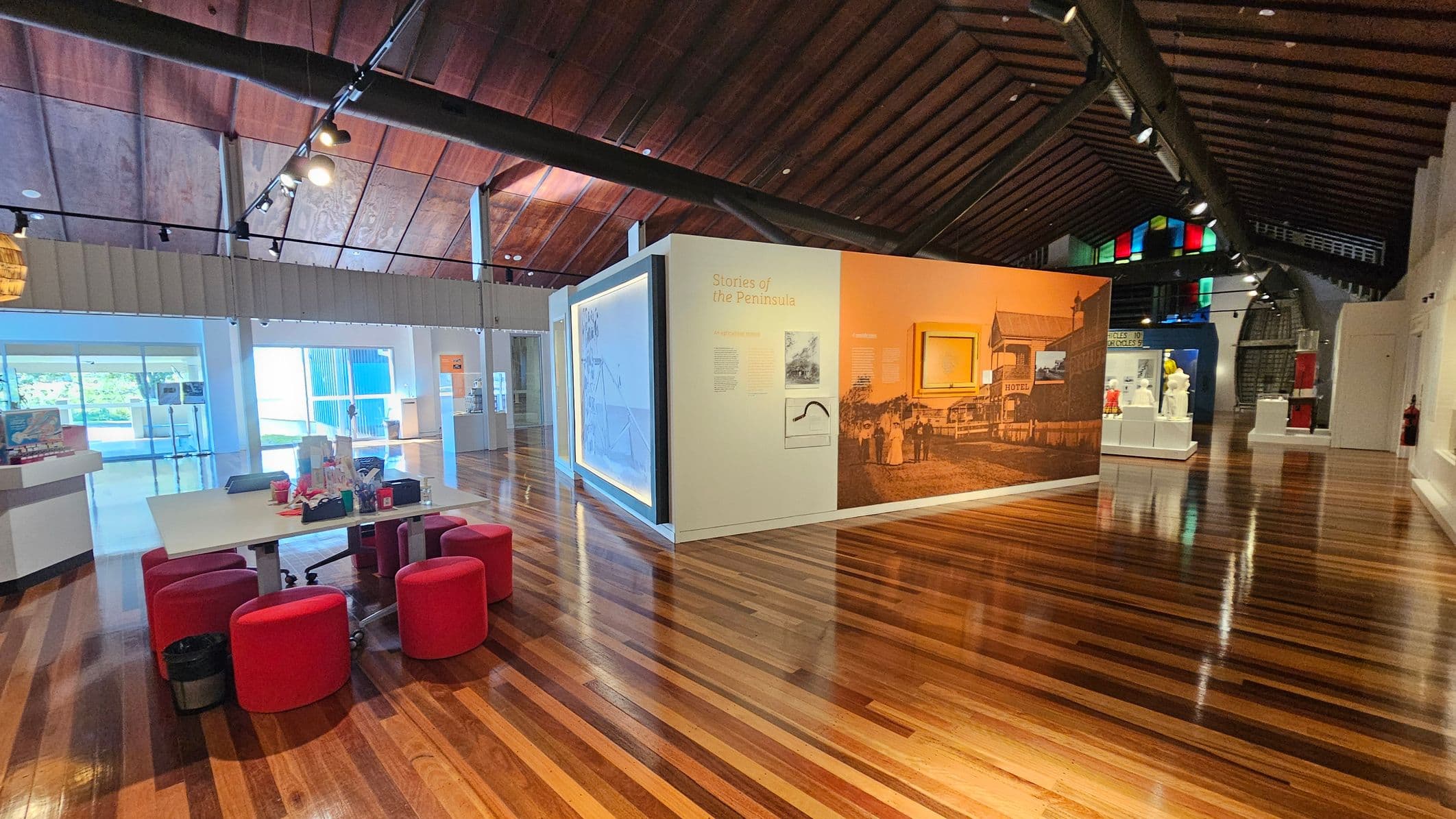 A spacious museum interior with polished wooden floors and a high, wooden ceiling. On the left, a table with red stools and various items on top. In the centre, a large display wall titled "Stories of the Peninsula" with historical images and text. The right side features more exhibits, including sculptures on pedestals. The background shows large windows allowing natural light and colourful panels near the ceiling.