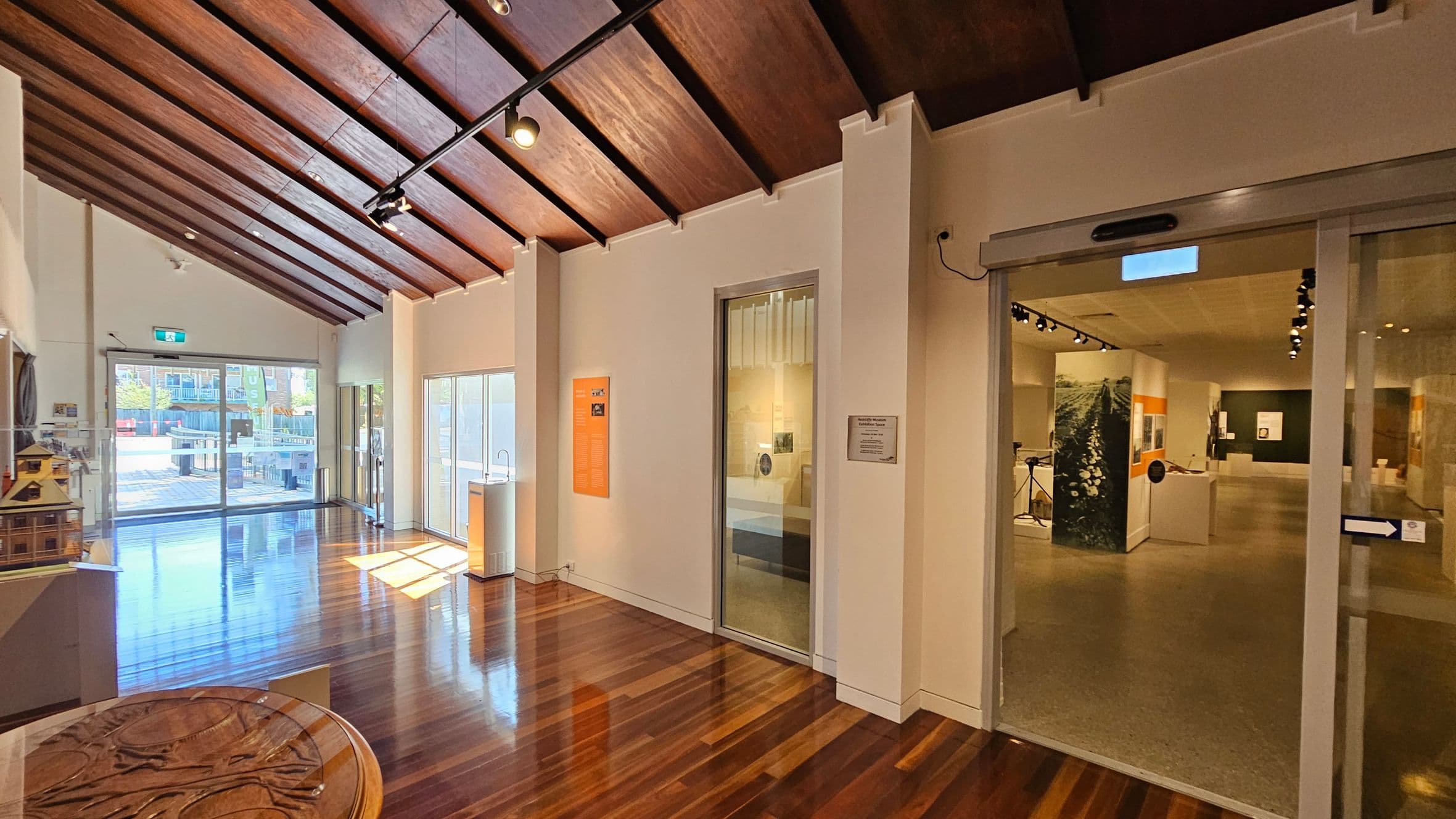 A museum interior with a wooden floor and a sloped wooden ceiling. On the left, there are large glass doors leading outside, allowing natural light to flood in. A small wooden model structure is visible on a table. In the centre, an orange informational panel is mounted on the wall. On the right, a glass sliding door opens into another exhibition space with displays and spotlights. The walls are light-coloured, and the overall atmosphere is bright and inviting.