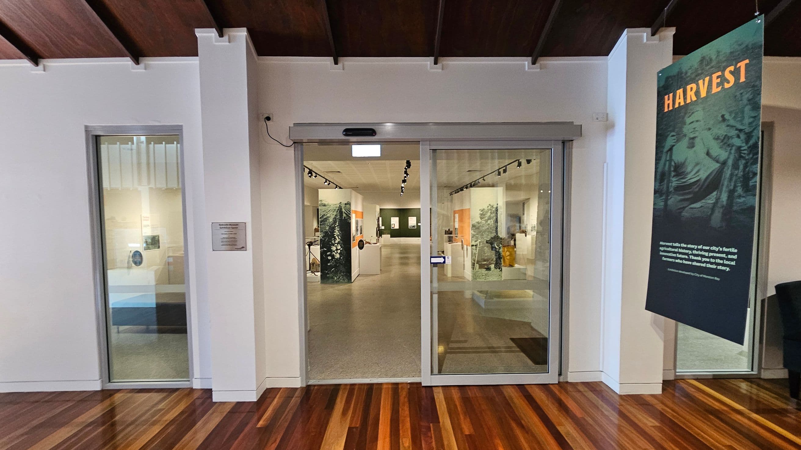 A glass entrance with sliding doors leads into a well-lit exhibition space. On the left, a window displays part of the exhibit. On the right, a large vertical banner reads "HARVEST" with additional text below. The floor is made of polished wood, and the ceiling features dark wooden beams. Inside, the exhibition area has displays and artwork on the walls, with track lighting overhead.