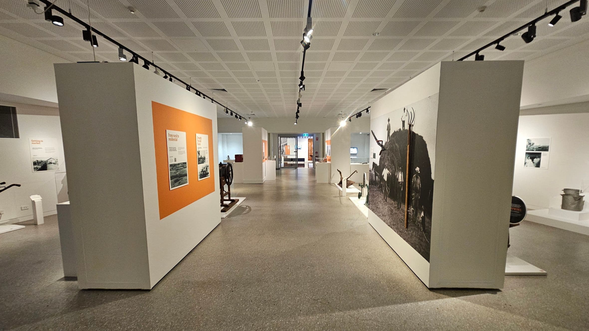 A museum interior with a wide corridor. On the left, a wall features an orange panel with text and images. On the right, a large black-and-white photograph of people with farming tools is displayed. The floor is grey and speckled, and the ceiling is white with a grid pattern, featuring track lighting. In the background, there are more exhibits and a glass exit door.