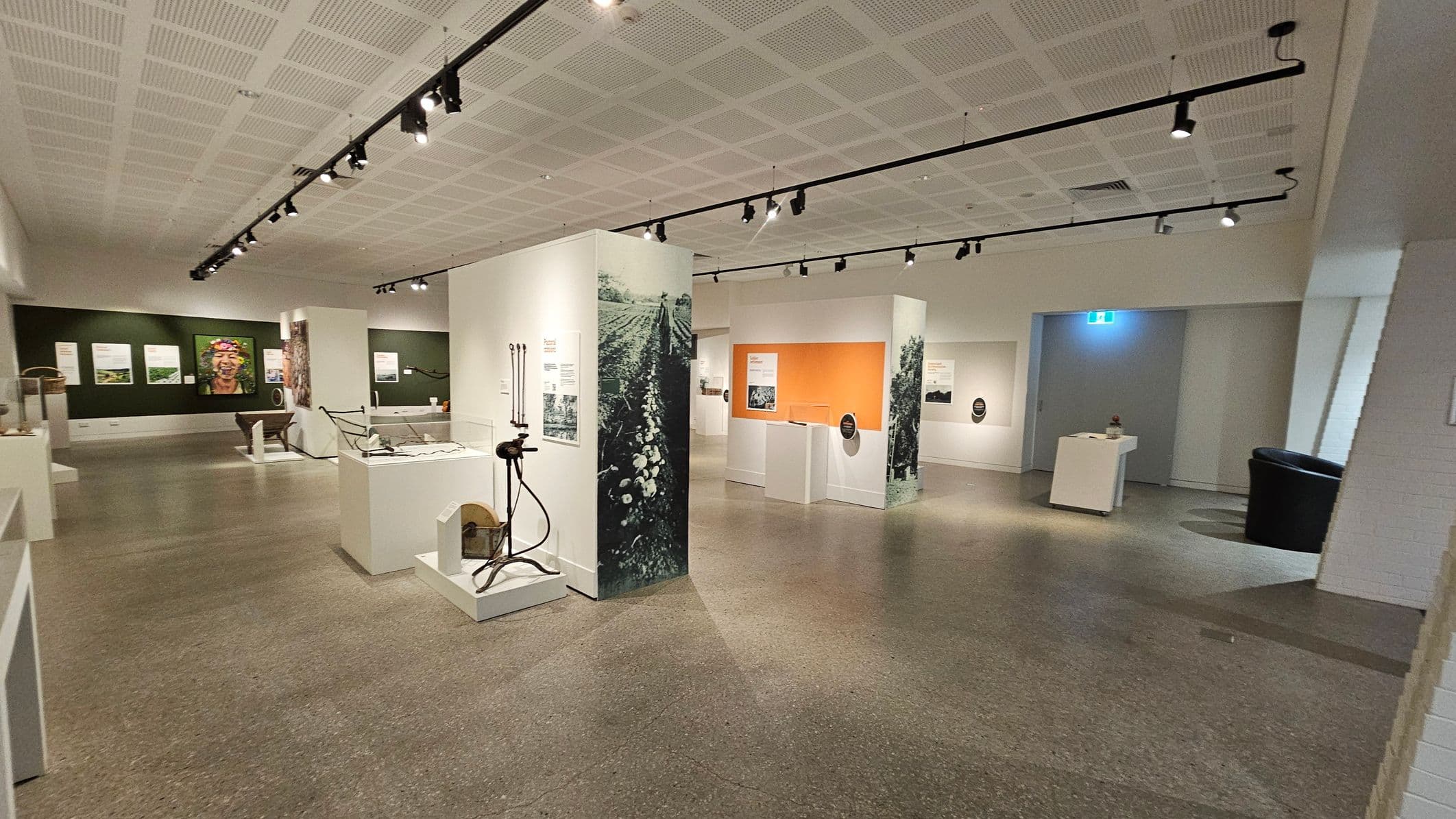 A spacious gallery with a grey speckled floor and a white perforated ceiling featuring track lighting. In the centre, there are display panels with agricultural images and tools. On the left, a green wall displays a large portrait and informational panels. To the right, an orange panel is visible with more displays. A small table and a black chair are near the back wall, which has a blue-lit exit sign.