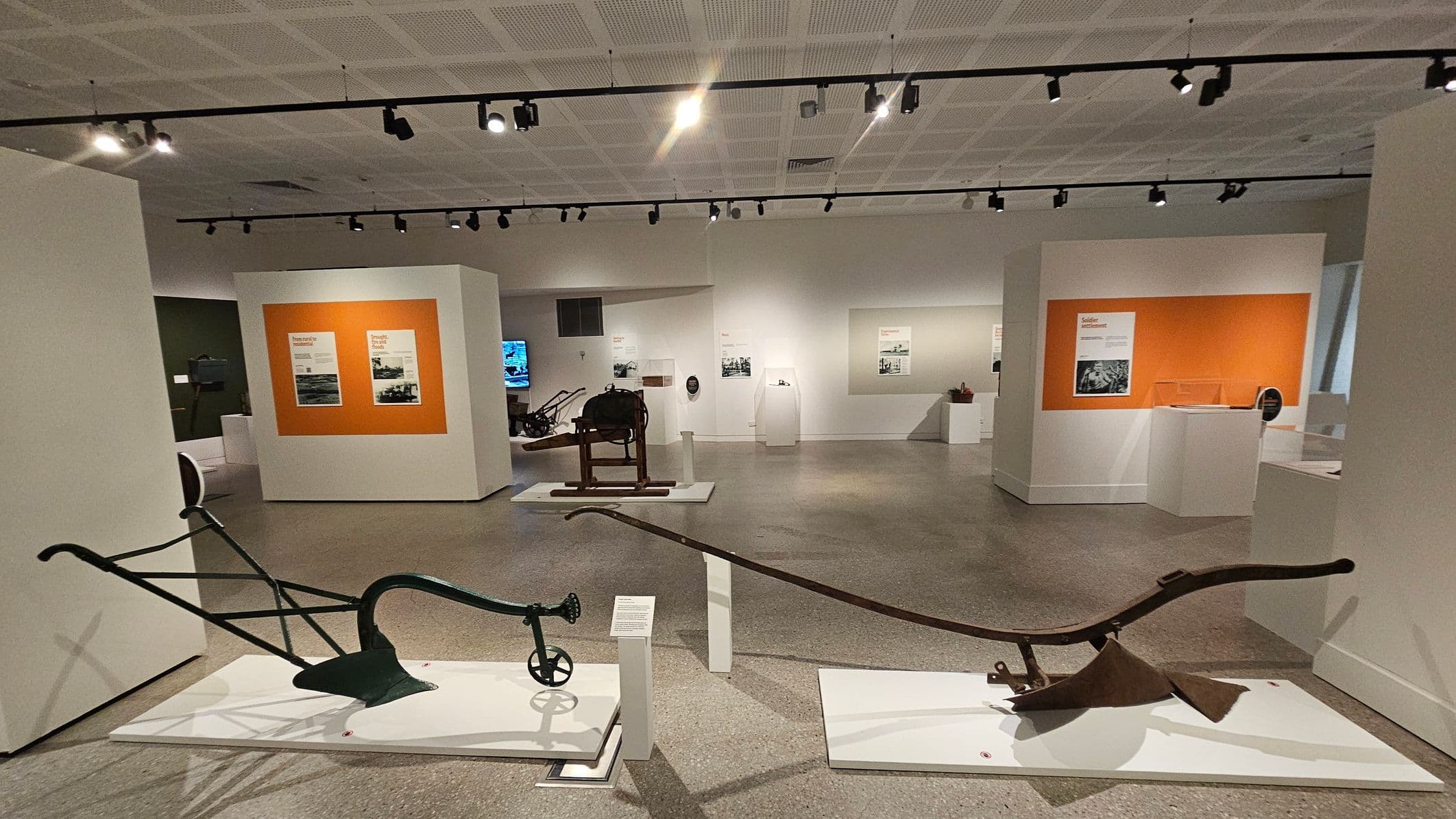 A museum exhibit with agricultural tools displayed on white platforms in the foreground. The left tool has a curved handle and a small wheel, while the right tool has a long, straight handle. The background features white walls with orange panels displaying text and images. Several other exhibits are visible, including a wooden piece on a stand. The ceiling is fitted with track lighting, and the floor is a smooth, grey surface.
