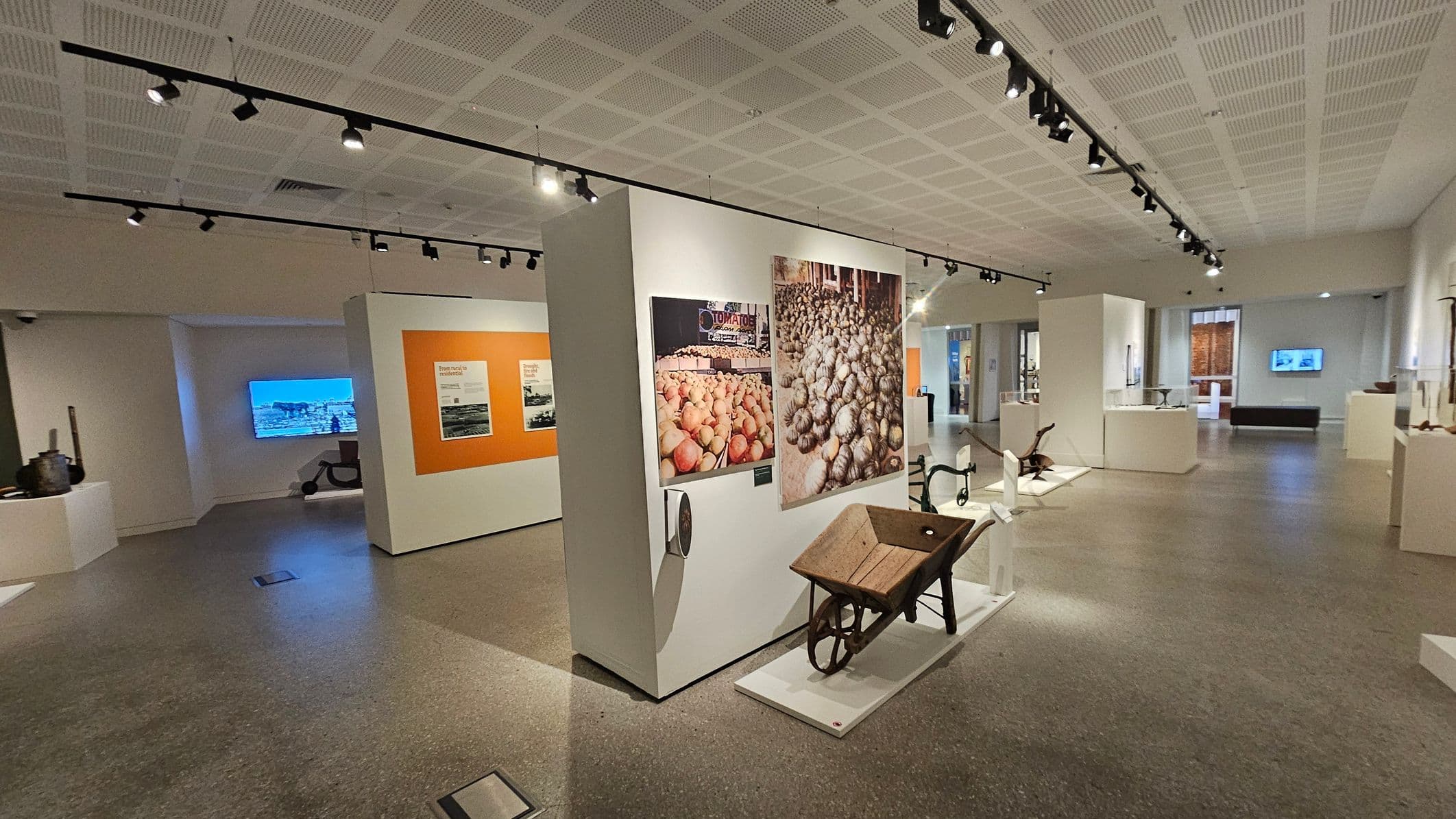 A museum interior with a polished grey floor and a white perforated ceiling with track lighting. In the centre, a display features a wooden wheelbarrow and large photographs of produce. To the left, an orange panel with text and images is visible. In the background, a screen displays a blue image, and various artefacts are arranged on white platforms. The space is open and well-lit, with exhibits spread throughout.