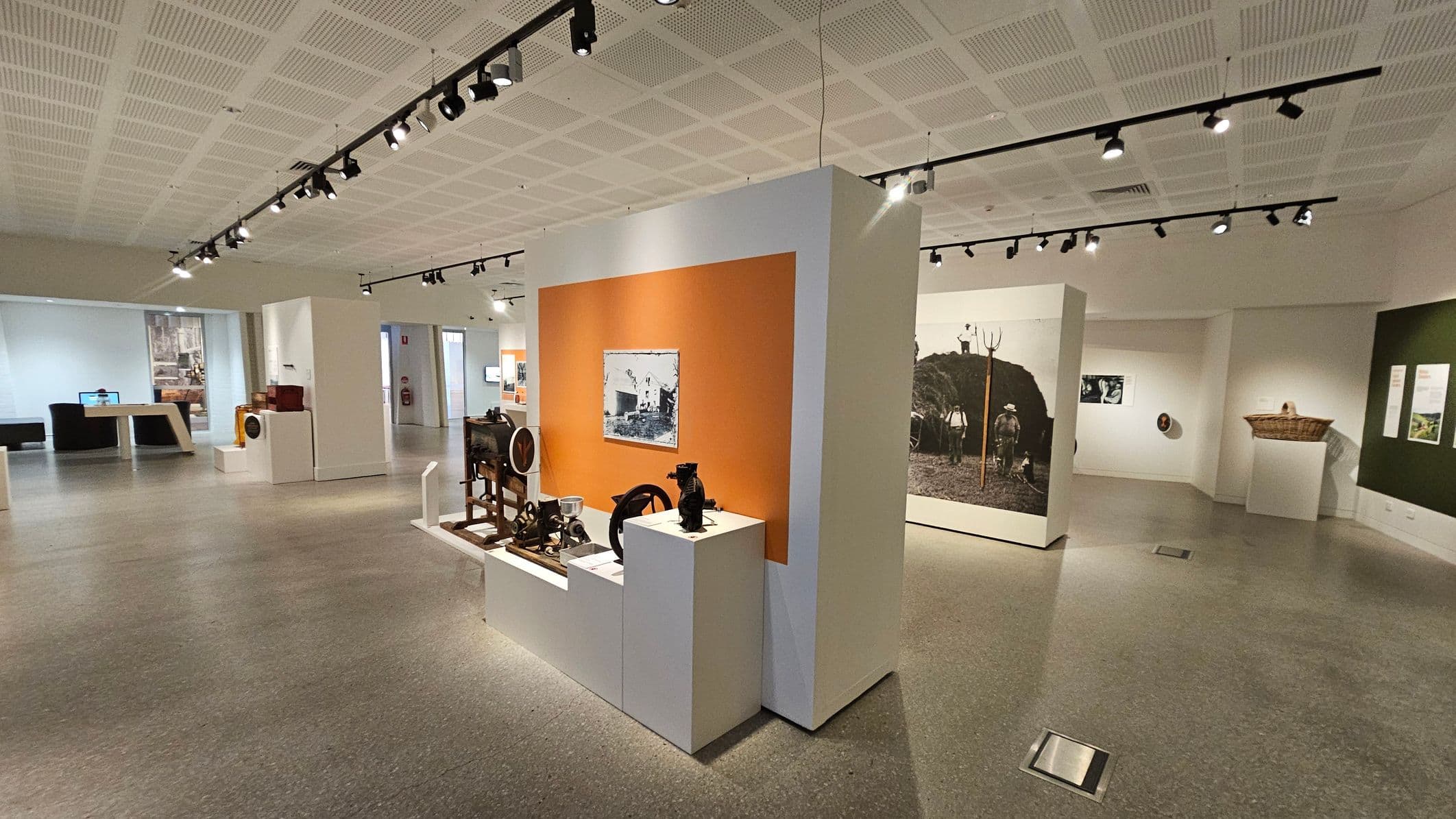 A spacious exhibition room with a grey speckled floor and a white, panelled ceiling with track lighting. In the centre, a display features various historical artefacts on white pedestals against an orange backdrop. To the right, a large black-and-white photograph of people with a pitchfork is mounted on a wall. On the left, a large window and a table with a computer screen are visible. A basket is mounted on the right wall, which is painted green.