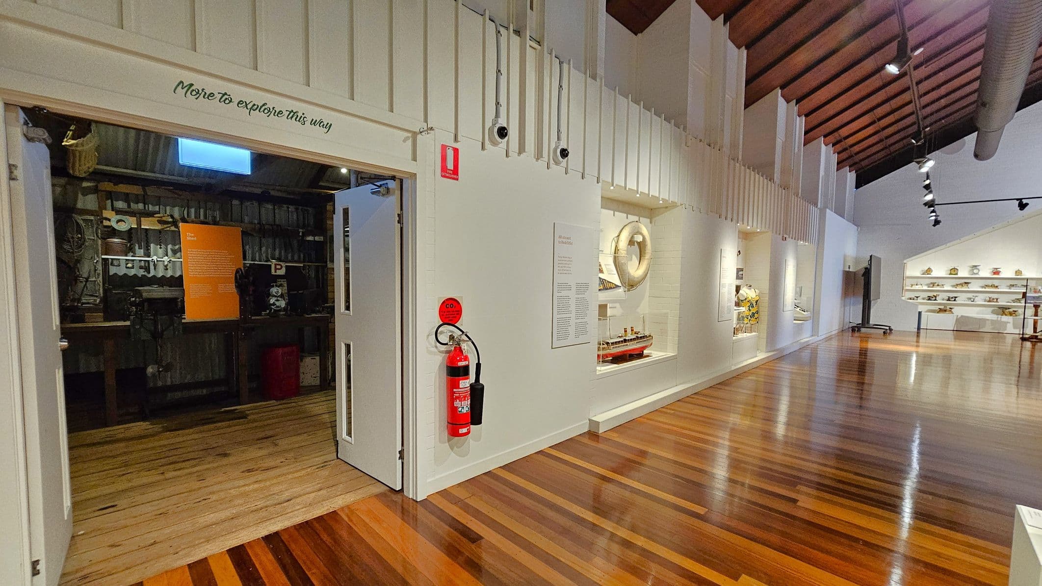 A museum interior with a wooden floor and a high, wooden-beamed ceiling. On the left, an open doorway leads to a room with tools and an orange information board. Above the door, a sign reads "More to explore this way." A fire extinguisher is mounted on the wall beside the door. On the right, display cases showcase various artefacts, including a model ship and a life preserver. Track lighting is installed on the ceiling, illuminating the exhibits.