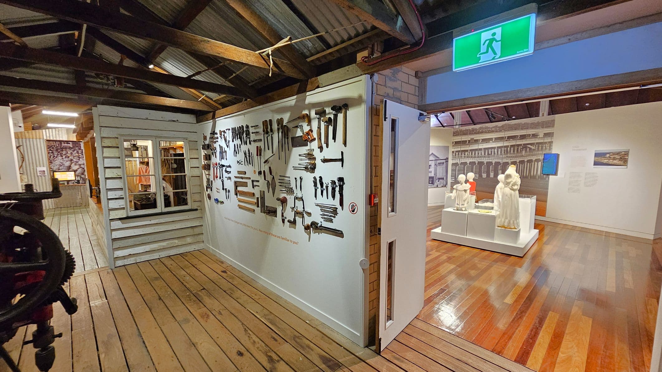A museum interior with wooden floors and a corrugated metal ceiling supported by wooden beams. On the left, a white wooden structure with a window displays various tools on the wall. In the centre, an open doorway leads to another room with mannequins dressed in historical clothing on a white platform. The background features a large black-and-white photograph of a building. An illuminated green exit sign is above the doorway.