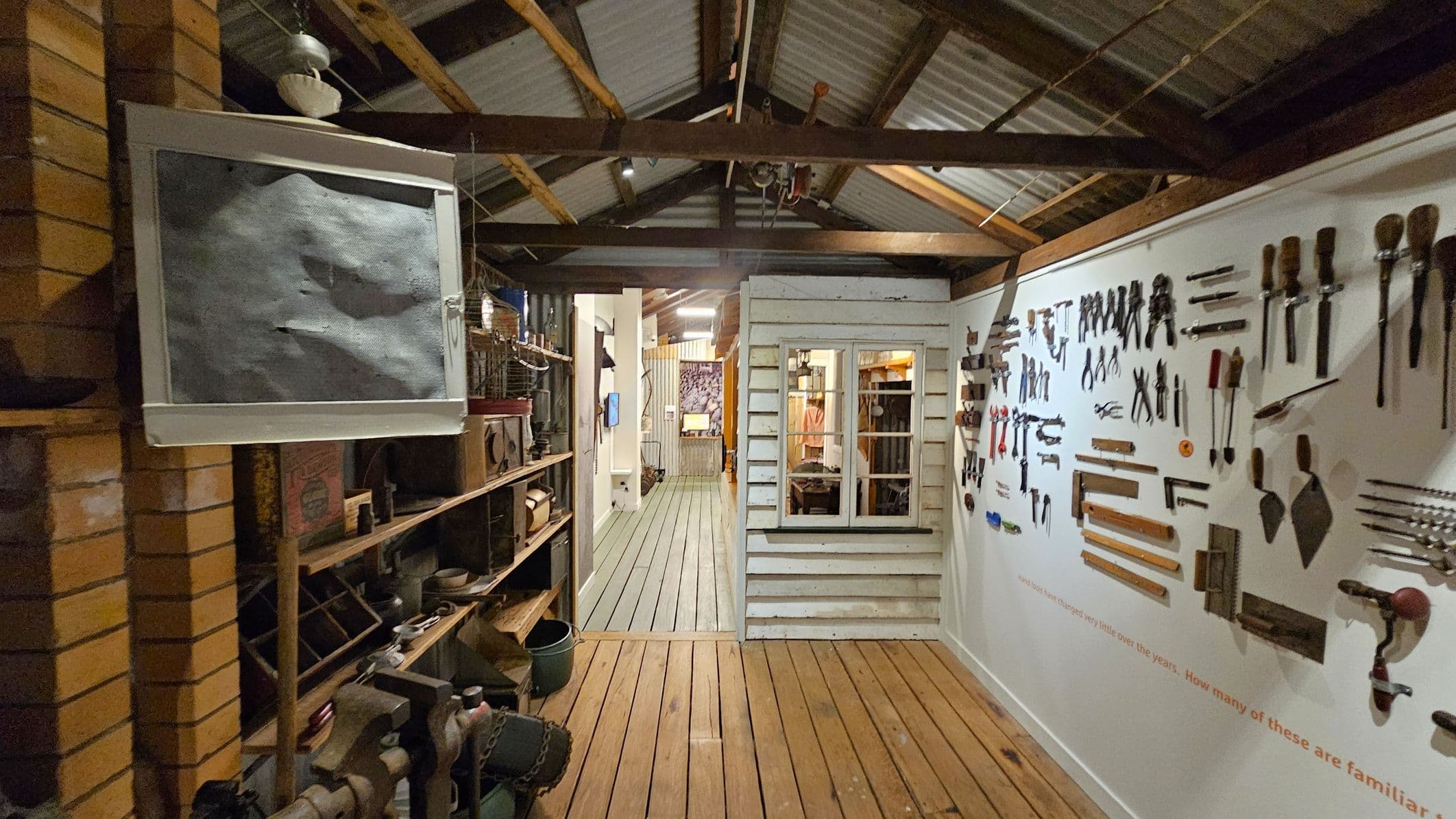 A rustic interior with a wooden floor and exposed beam ceiling. On the left, a brick wall with shelves holding various vintage items. On the right, a white wall displays an array of tools neatly arranged. In the centre, a small structure resembling a house with a window and door, leading to a corridor with a green wooden floor. The background shows more rooms with additional items and displays.