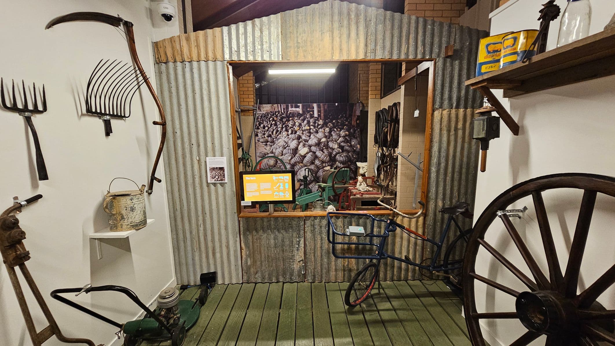 A museum exhibit featuring vintage farming tools. On the left, a scythe, pitchforks, and a watering can are displayed on a white wall. A green lawnmower is on the floor. In the centre, a corrugated metal structure with an open window reveals a backdrop of pumpkins and various tools. An informational screen is visible. On the right, a large wooden wheel and a bicycle with a basket are displayed. The floor is wooden, and the ceiling is not visible.