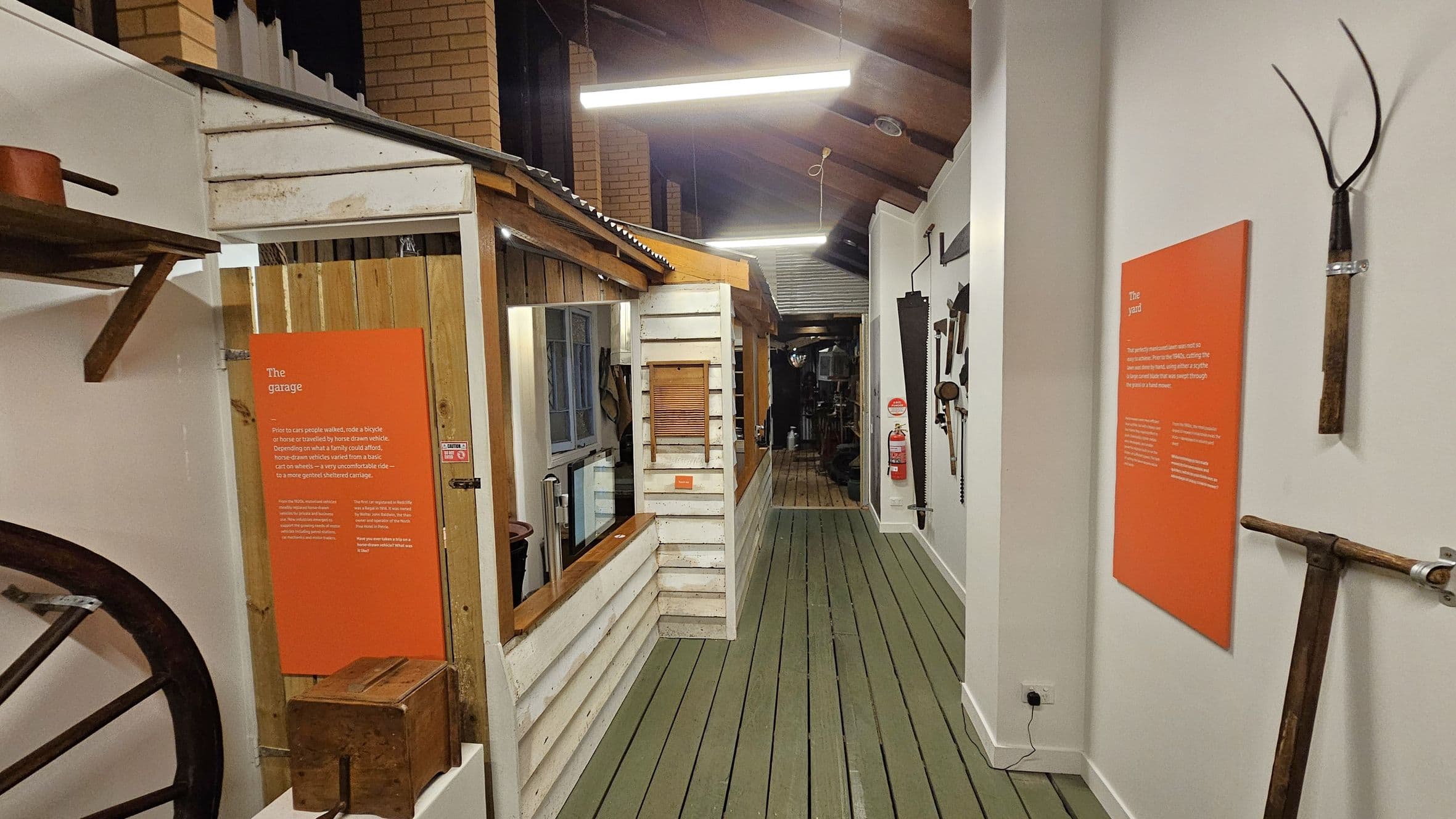 A narrow indoor corridor with a wooden floor and a high ceiling with exposed beams and fluorescent lights. On the left, there's a wooden structure resembling a small house or garage with a corrugated metal roof. An orange informational sign is mounted on the wall. On the right, various tools are displayed on a white wall, along with another orange sign. A fire extinguisher is visible further down the corridor.