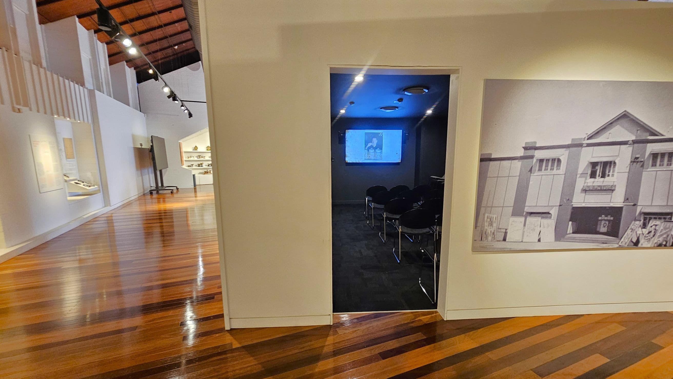 A room with polished wooden floors and a high ceiling with exposed beams. On the left, a corridor with display cases and a track of spotlights. In the centre, an open doorway leads to a dark room with chairs facing a screen showing a projected image. On the right, a large black-and-white photograph of a building is mounted on the wall.