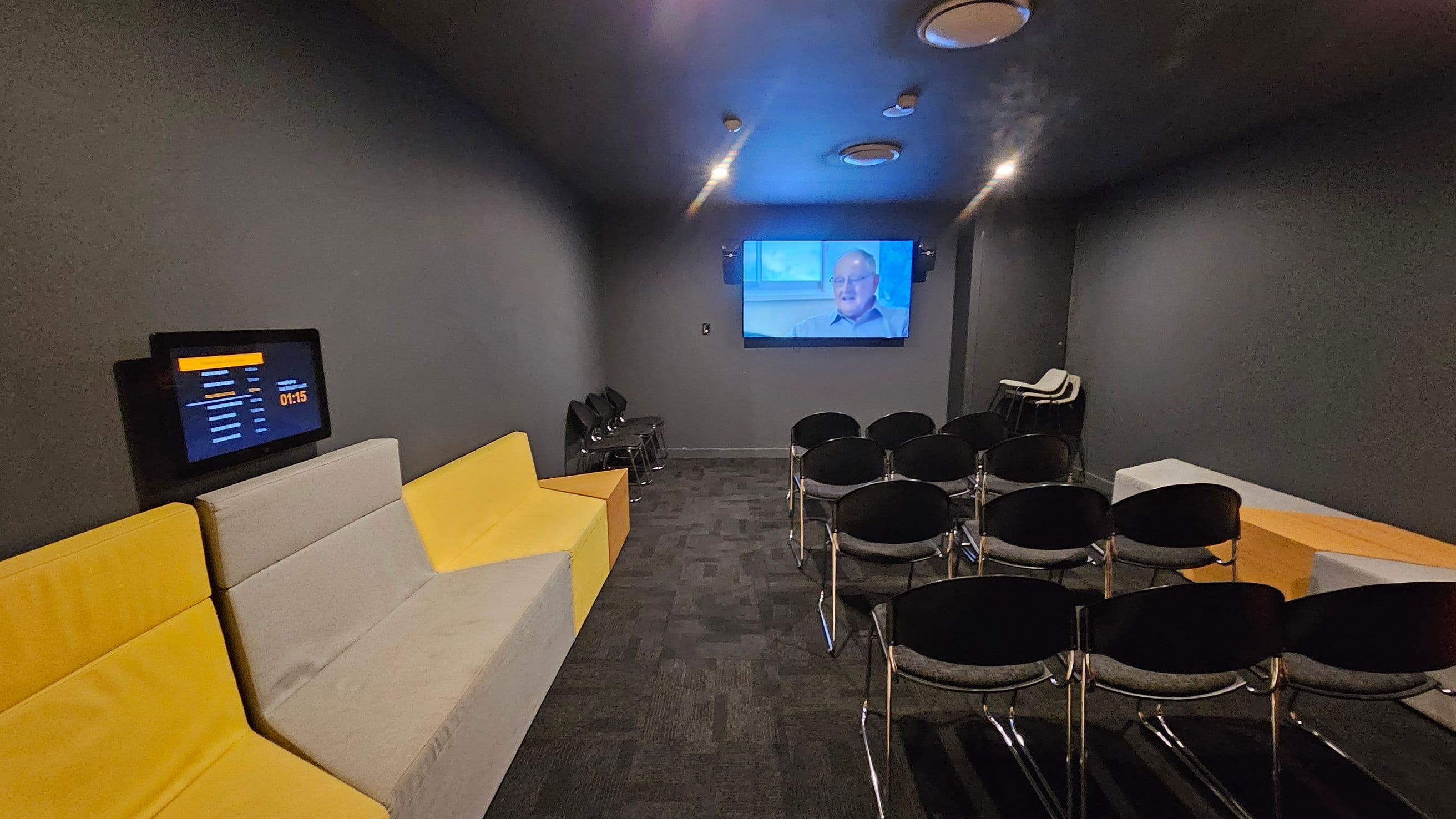 A small, dimly lit room with a grey ceiling and walls. On the left, there is a yellow and grey sofa with a wall-mounted screen displaying a countdown timer. In the centre, several rows of black chairs face a large screen on the far wall, showing a person speaking. On the right, a stack of additional chairs is visible. The floor is carpeted in a dark, patterned design.