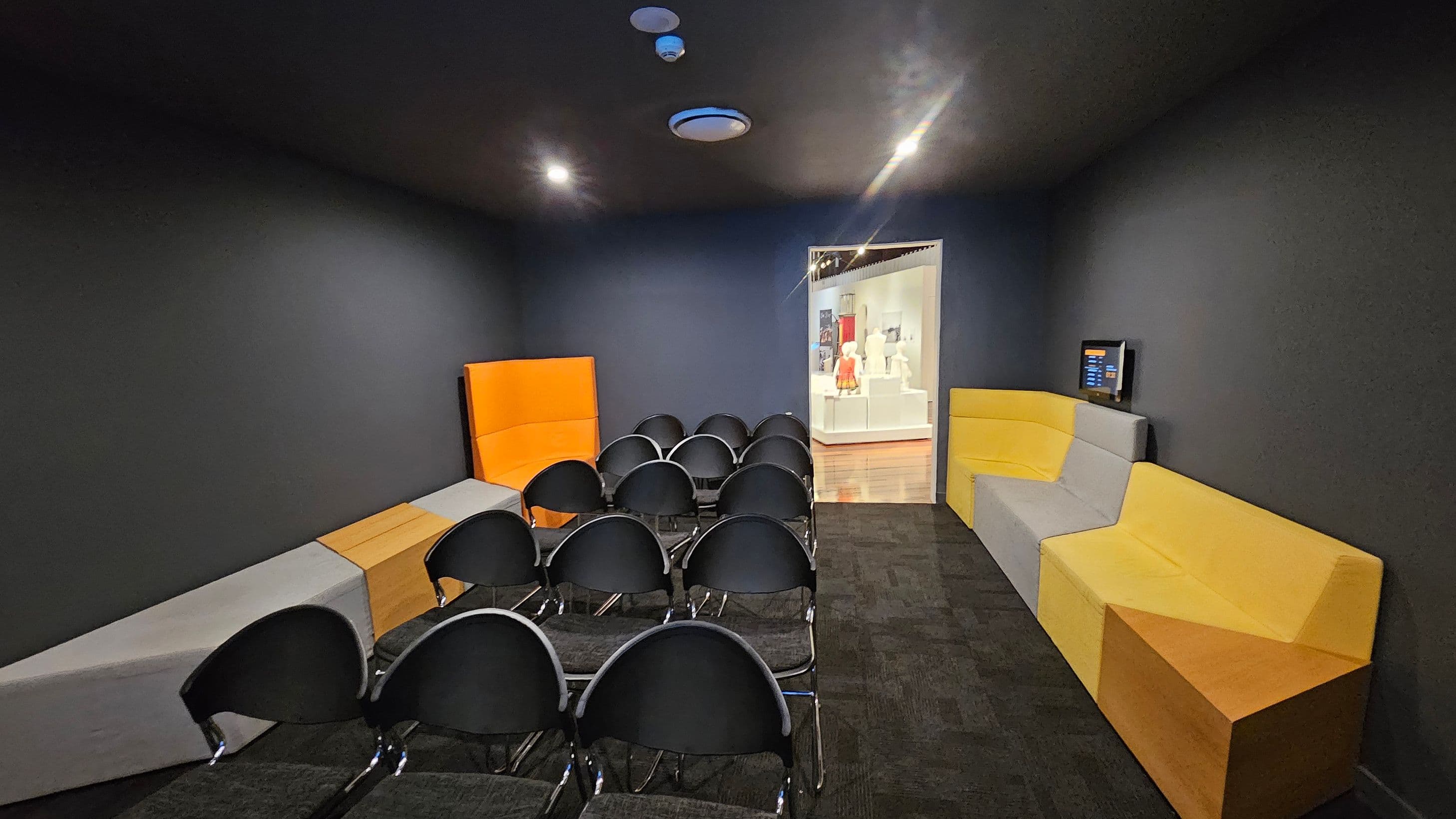 A small room with dark walls and ceiling, featuring rows of black chairs in the centre. On the left, there are angular benches in grey and orange. On the right, similar benches in yellow and grey are present, with a small screen mounted on the wall. At the back, an open doorway leads to a brightly lit area displaying mannequins. The floor is carpeted in dark tones.