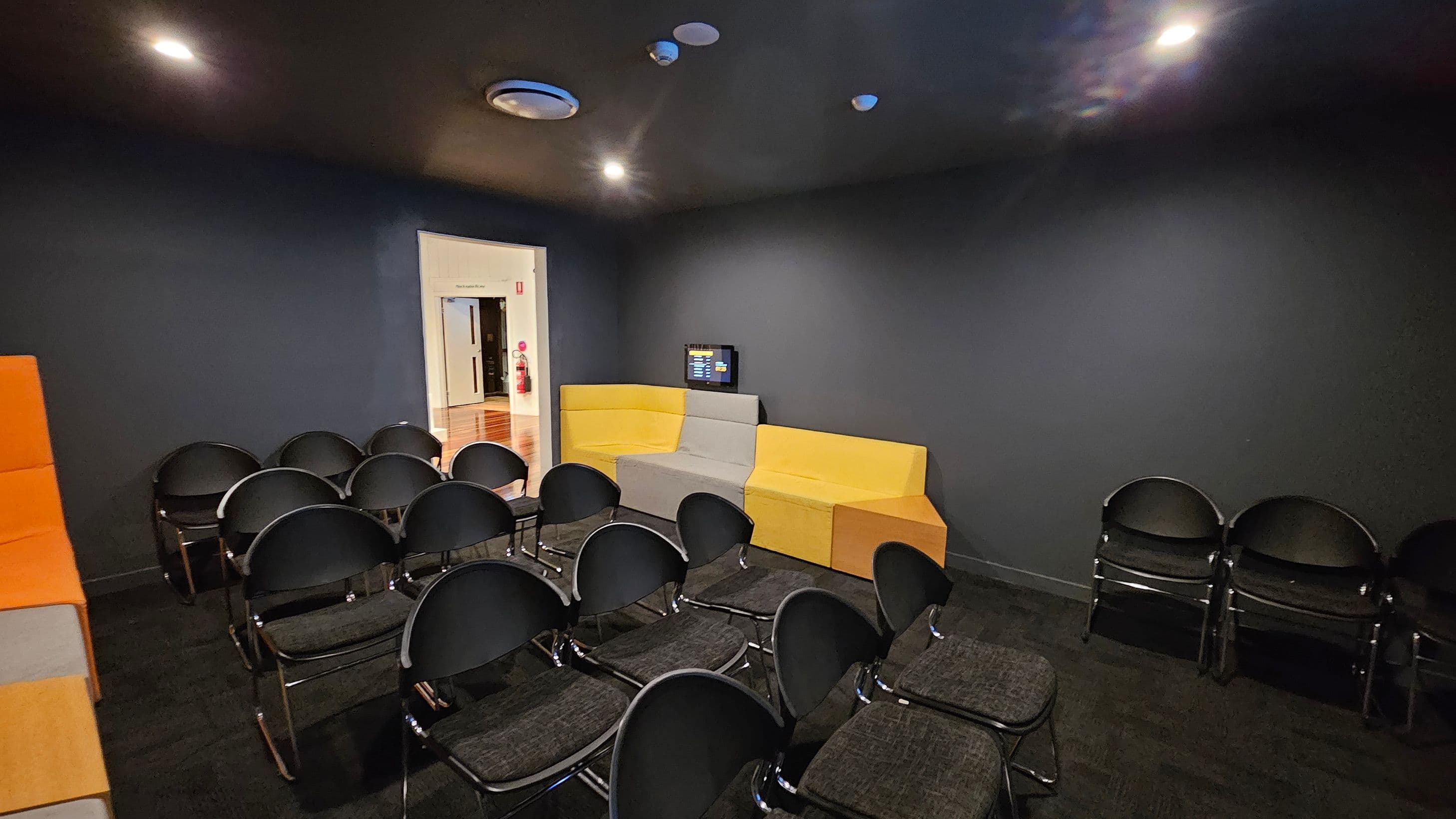 A small room with dark walls and ceiling, featuring a cluster of black chairs arranged in rows on a dark carpeted floor. On the right, there are yellow and grey modular sofas against the wall, with a small screen mounted above them. In the background, an open doorway leads to a hallway with wooden flooring and a visible fire extinguisher. The ceiling has recessed lighting and a circular vent.