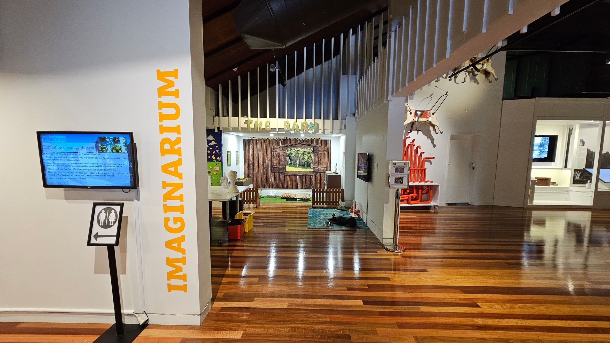 A room with wooden flooring and a high ceiling. On the left, a wall with the word "IMAGINARIUM" in yellow and a mounted screen displaying information. Below the screen, a sign with a restroom symbol and an arrow. In the centre, an area labelled "THE BARN" with a rustic wooden backdrop and small fence. On the right, a display with red pipes and a white room with exhibits. Hanging from the ceiling are animal figures.