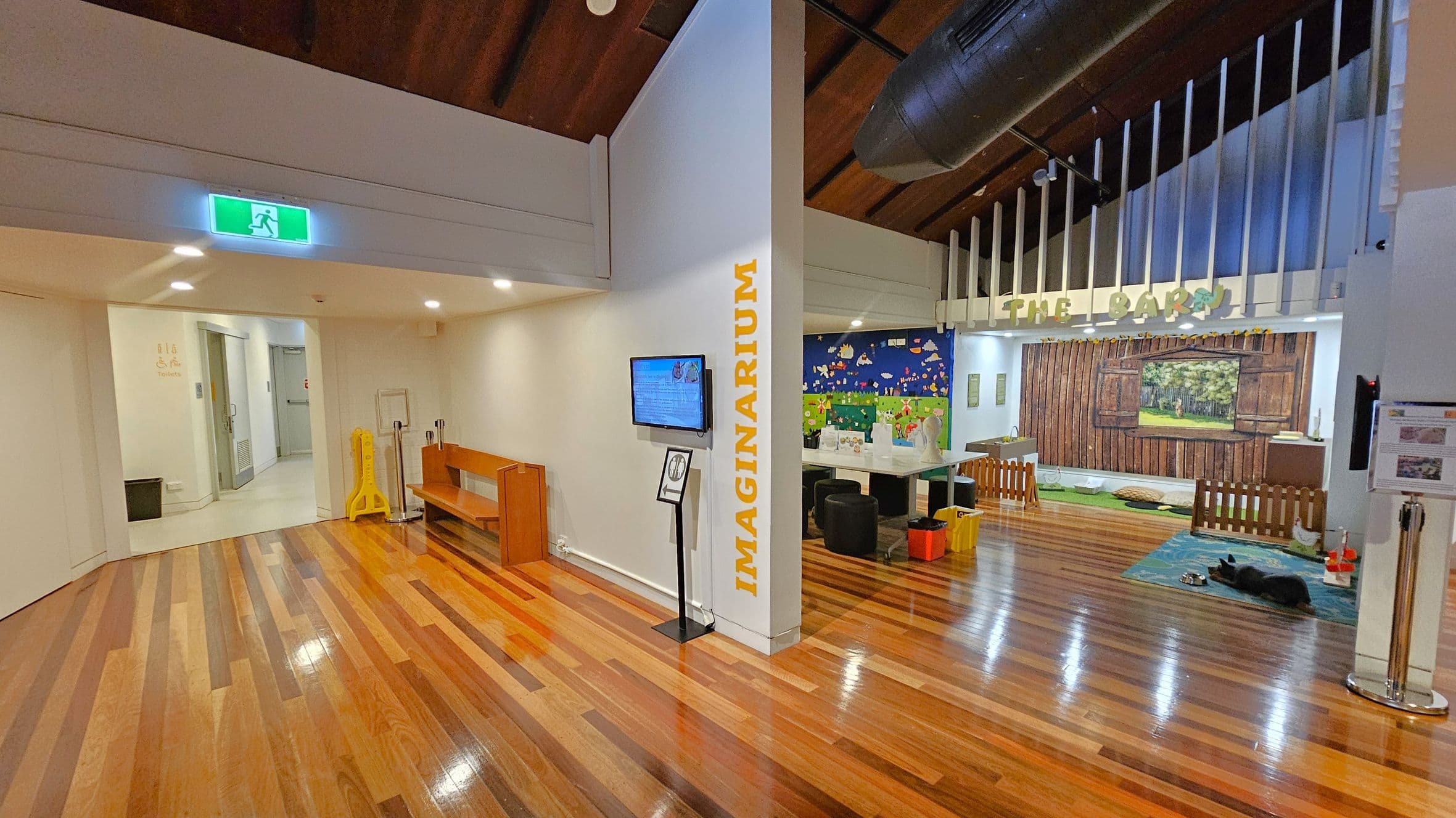A room with polished wooden floors and a high wooden ceiling. On the left, there's a corridor leading to toilets, marked by a green exit sign. A wooden bench and a yellow caution sign are nearby. In the centre, a wall with "IMAGINARIUM" written vertically and a mounted screen. On the right, an area called "THE BARN" with a barn-themed backdrop, a small fenced area, and a rug with a dog lying on it. The ceiling has exposed beams and ductwork.