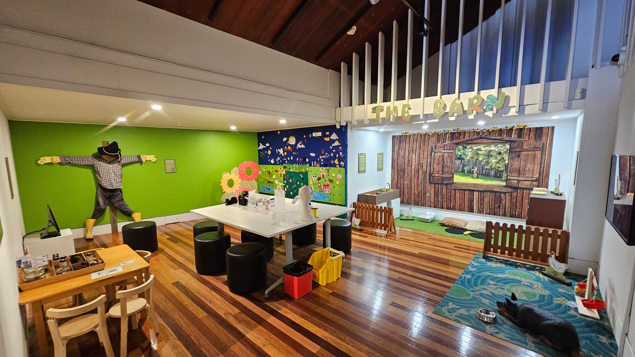 A room with a wooden floor and a high ceiling. On the left, a green wall features a scarecrow figure. A small wooden table with chairs is nearby. In the centre, a large white table with black stools and colourful bins underneath. The right side has a mural of a barn with a wooden fence and a small play area with a rug and a toy dog. The back wall has a colourful farm-themed mural and a sign reading "THE BARN" above.
