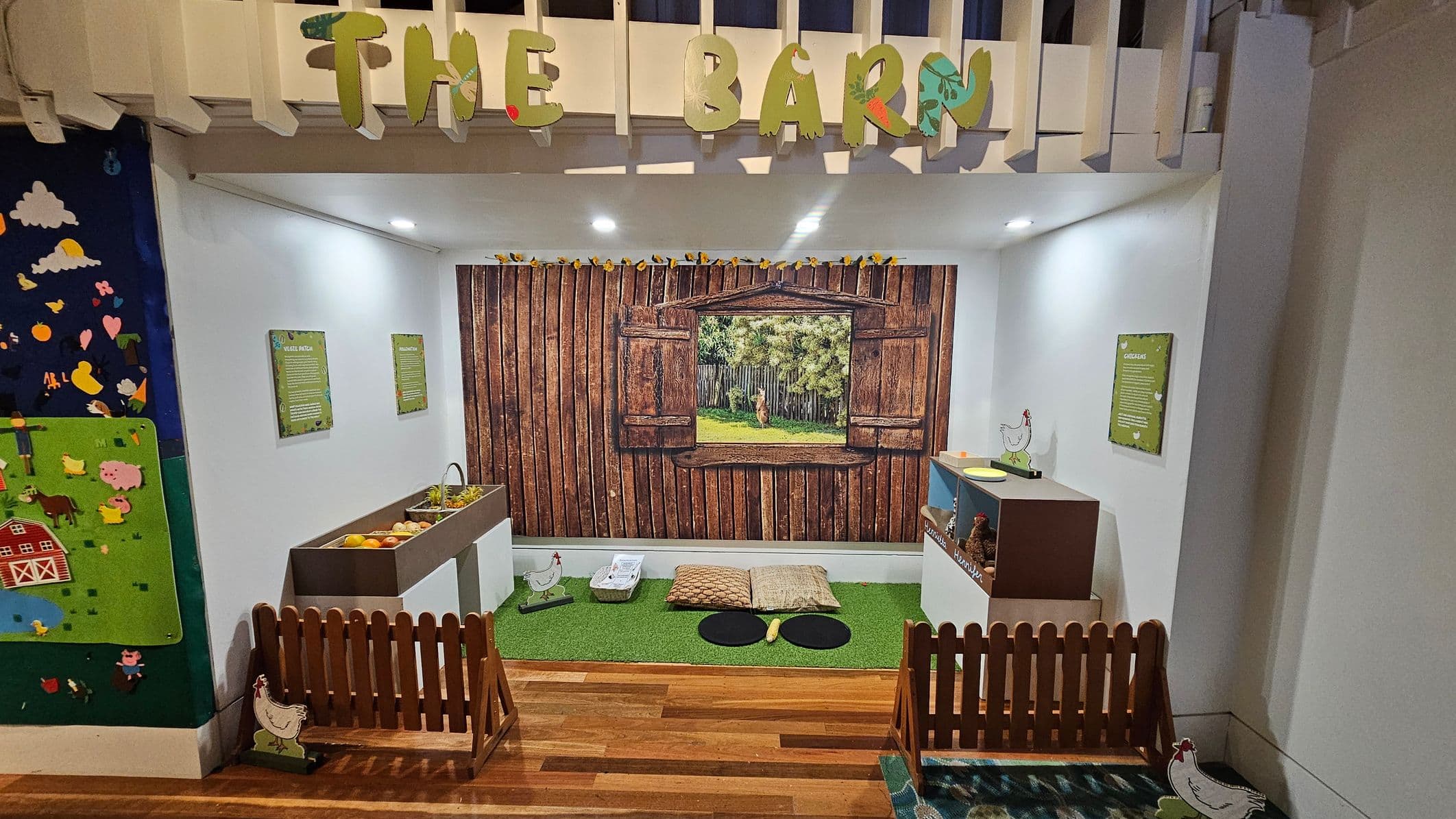A children's play area designed to resemble a barn. The background features a wooden wall with a window showing a farm scene. In the foreground, there are two small fences with chicken cutouts. On the left, a sink with toy fruits and vegetables. On the right, a display with toy chickens and eggs. The floor is wooden with a green mat in the centre, and the ceiling has a sign reading "THE BARN" with decorative letters. Walls have educational signs.
