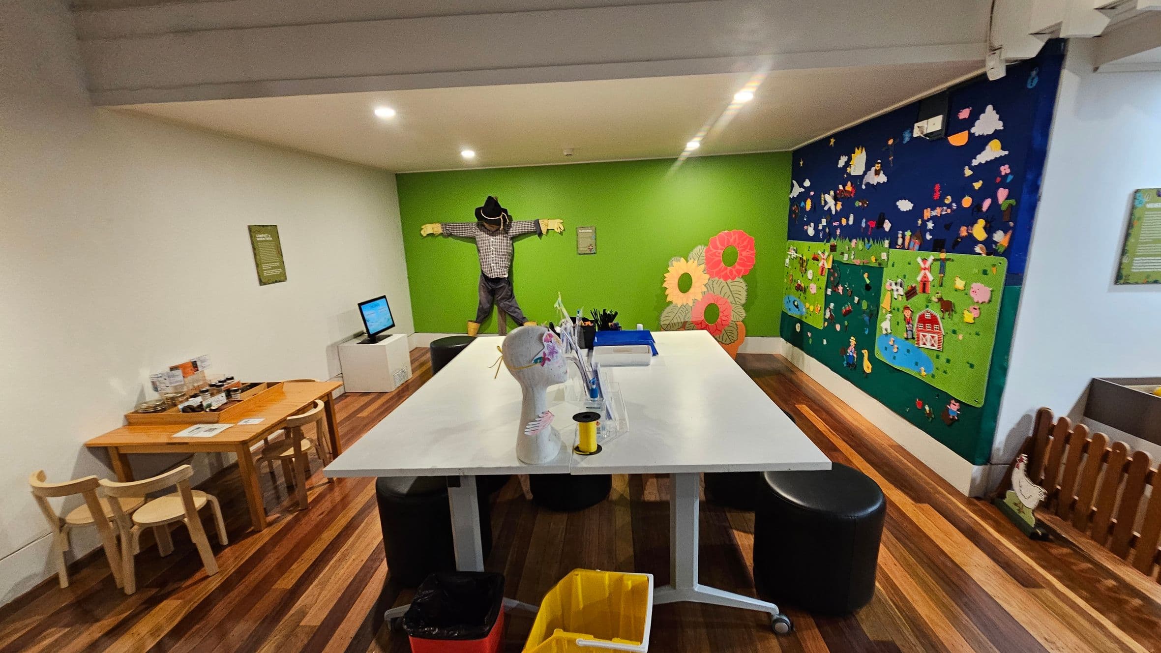 A room with wooden flooring and a white ceiling. In the centre, a large white table with various craft supplies and a mannequin head. A yellow bin is on the floor nearby. On the left, a small wooden table with chairs and a computer monitor. The back wall is green with a scarecrow and large flower decorations. The right wall features a colourful mural depicting a farm scene with animals and a barn.