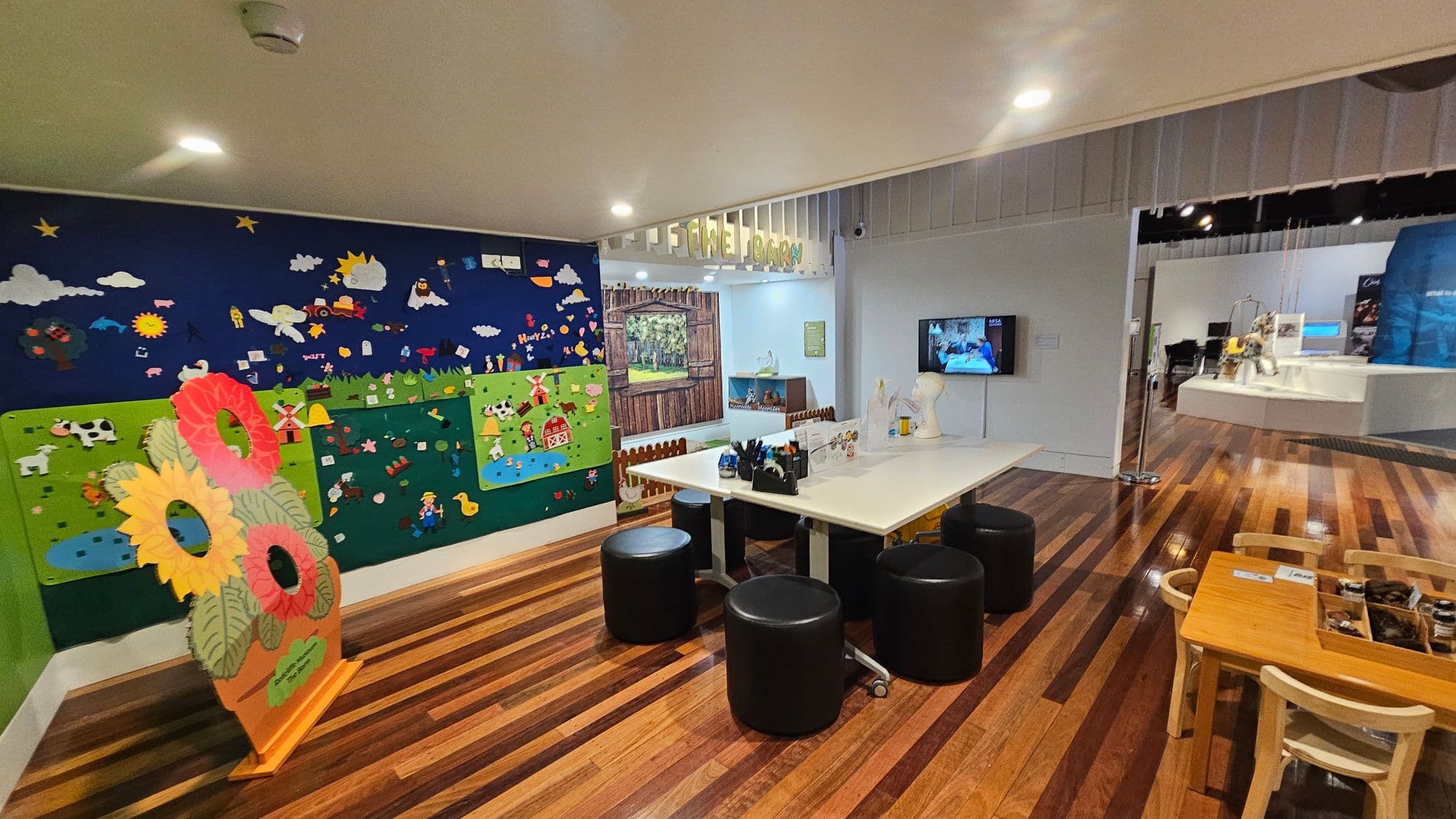 A children's play area with a colourful wall mural on the left, featuring farm animals and nature scenes. In the foreground, a stand with large flower cutouts. A white table with black stools is in the centre, surrounded by art supplies. On the right, a small wooden table with chairs and a display case. The floor is wooden, and the ceiling is white with recessed lighting. A TV screen is mounted on the wall, and a sign above reads "The Farm".