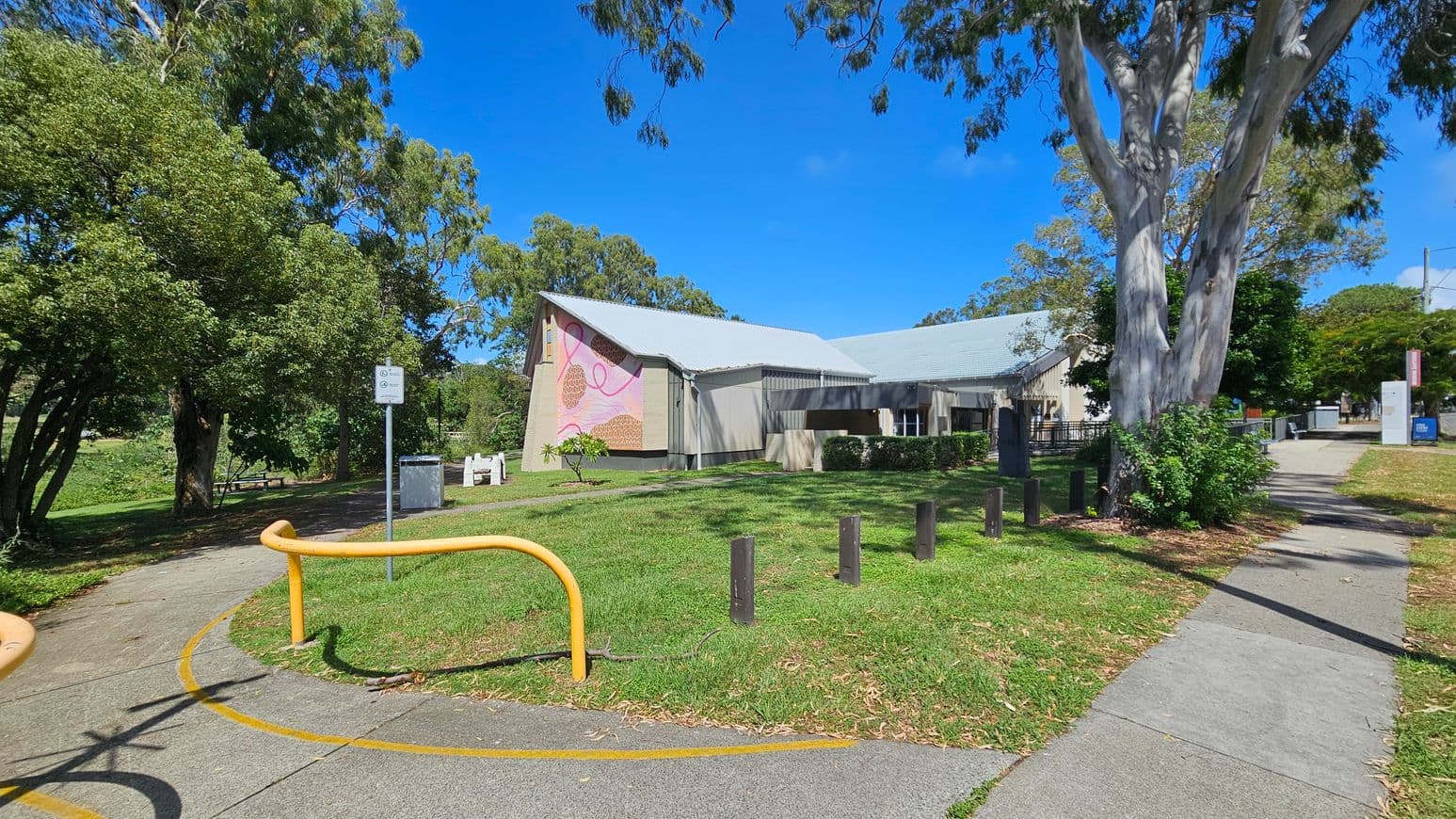 A building with a grey roof and a colourful mural on the side is situated in a grassy area. To the left, there are trees and a path with a yellow barrier. A signpost and a bin are nearby. On the right, a large tree stands next to a paved walkway leading towards the building. Bushes and small plants are scattered around, and there are wooden posts along the grass. The sky is clear and blue.