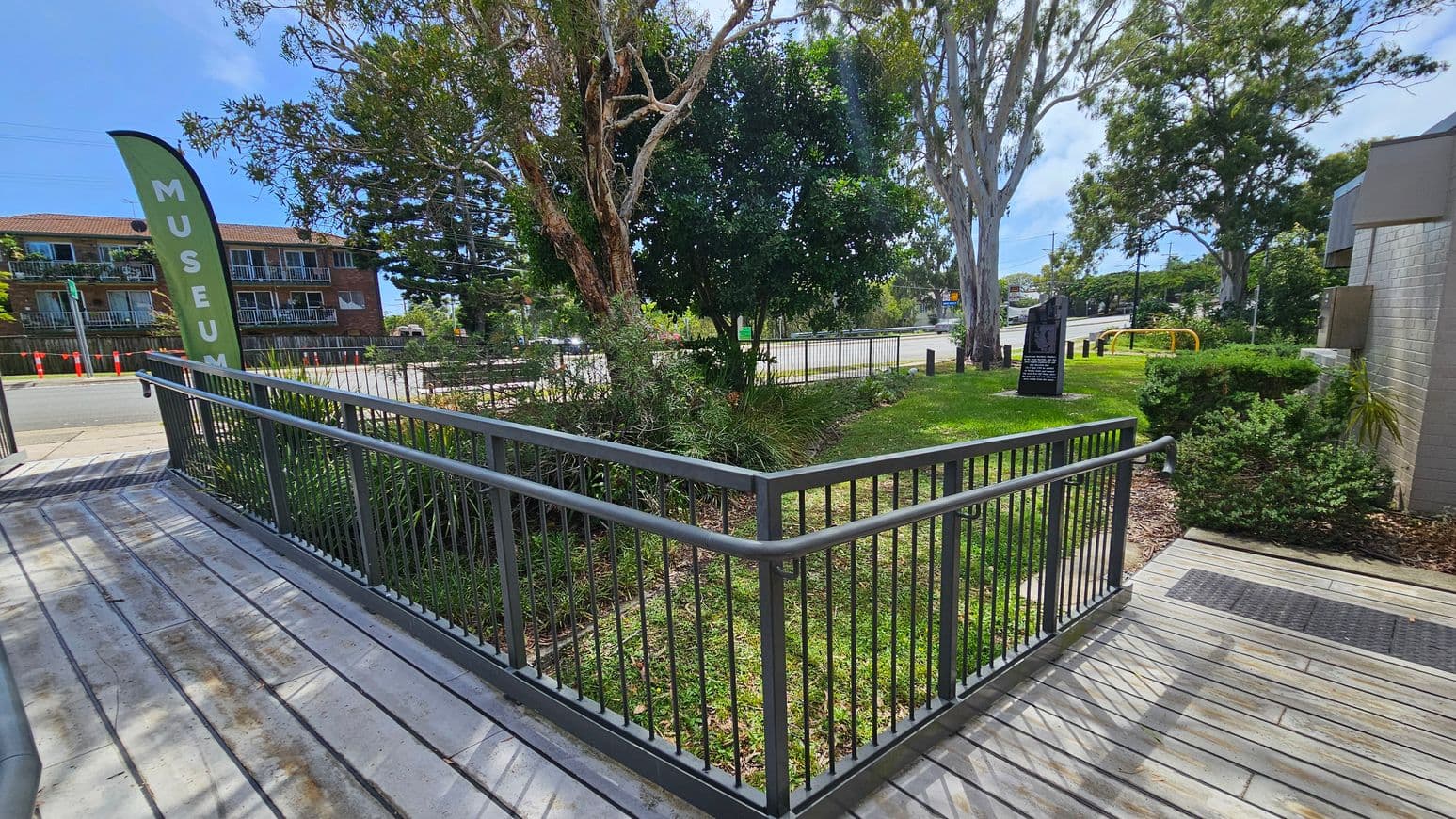 A wooden walkway with metal railings leads to a grassy area on the right. A tall green flag with the word "MUSEUM" is on the left. In the background, there are trees and a brick building with balconies. A black information sign is visible on the grass, and a yellow bike rack is near the road. The area is bordered by a street with orange traffic cones.