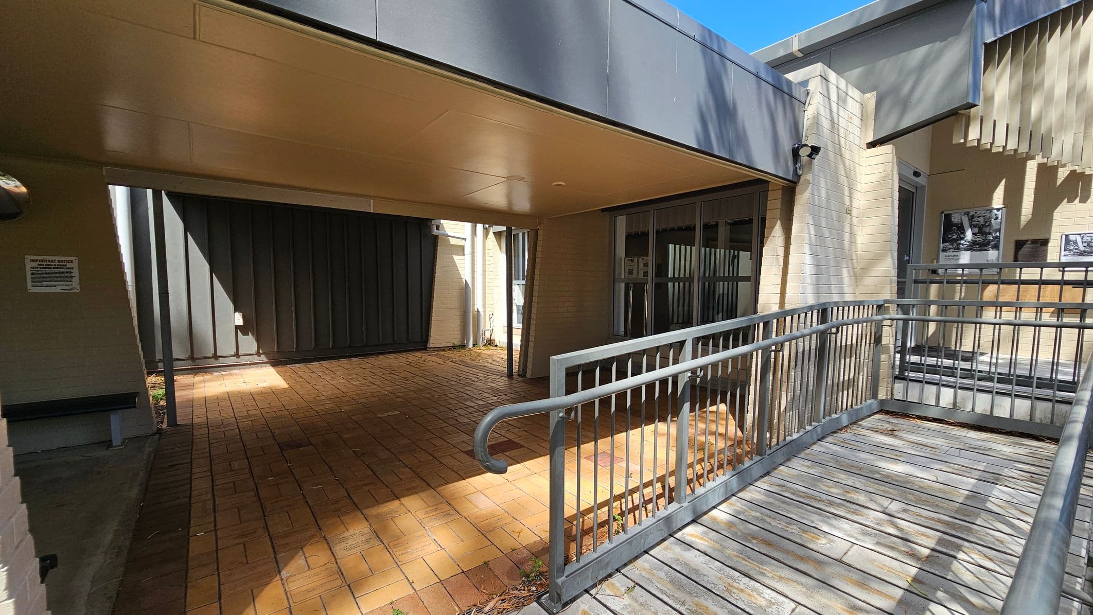 A covered walkway with a brick floor leads to a building entrance. On the right, a metal ramp with railings provides access to a door with glass panels. The building is constructed with light-coloured bricks and has a grey overhang. There are some posters or notices on the wall near the entrance. Shadows from nearby trees are cast on the walls and floor. A sign is visible on the left wall.