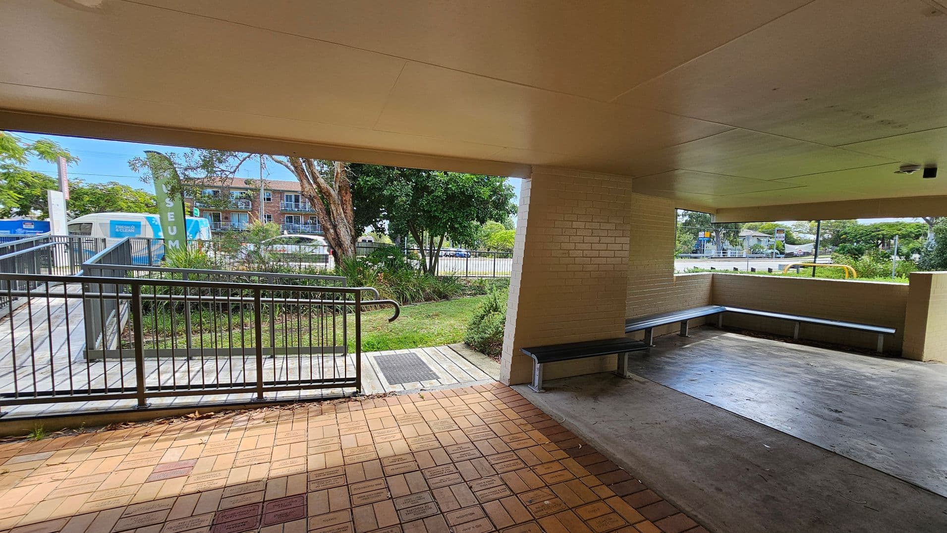 A covered outdoor area with a tiled floor and a ceiling. On the right, there is a corner bench with metal seating. The left side features a ramp with metal railings leading to a grassy area with trees. In the background, a building with balconies is visible, along with a blue and white vehicle. A green flag with partial text is near the ramp. The area is surrounded by greenery and a fence.
