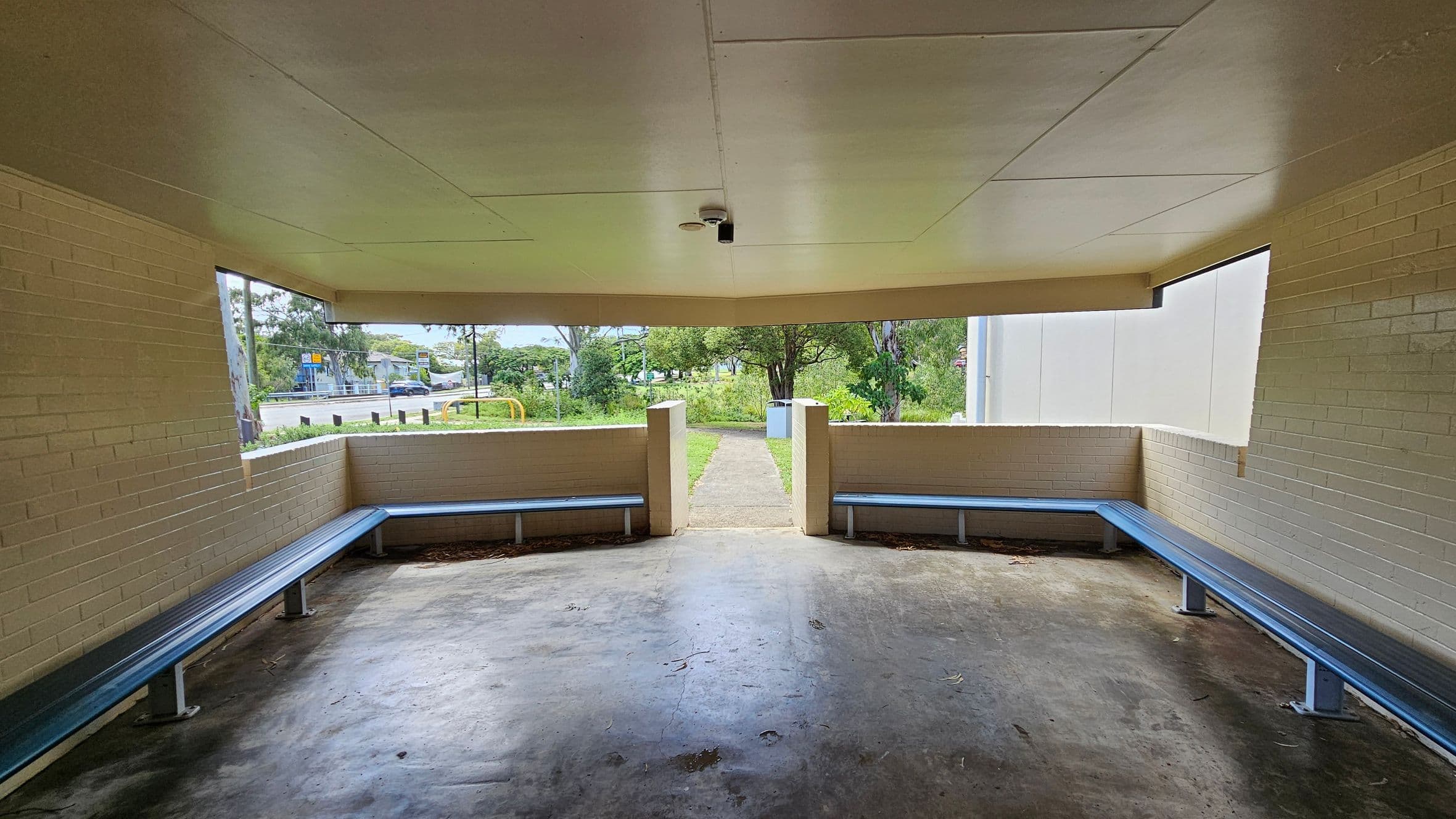 A sheltered area with beige brick walls and a flat ceiling. Two long blue benches are fixed along the left and right walls. The floor is concrete with some dirt and leaves scattered. The open front leads to a grassy area with trees and a path. A security camera is mounted on the ceiling. In the background, a road and some buildings are visible through the open sides.