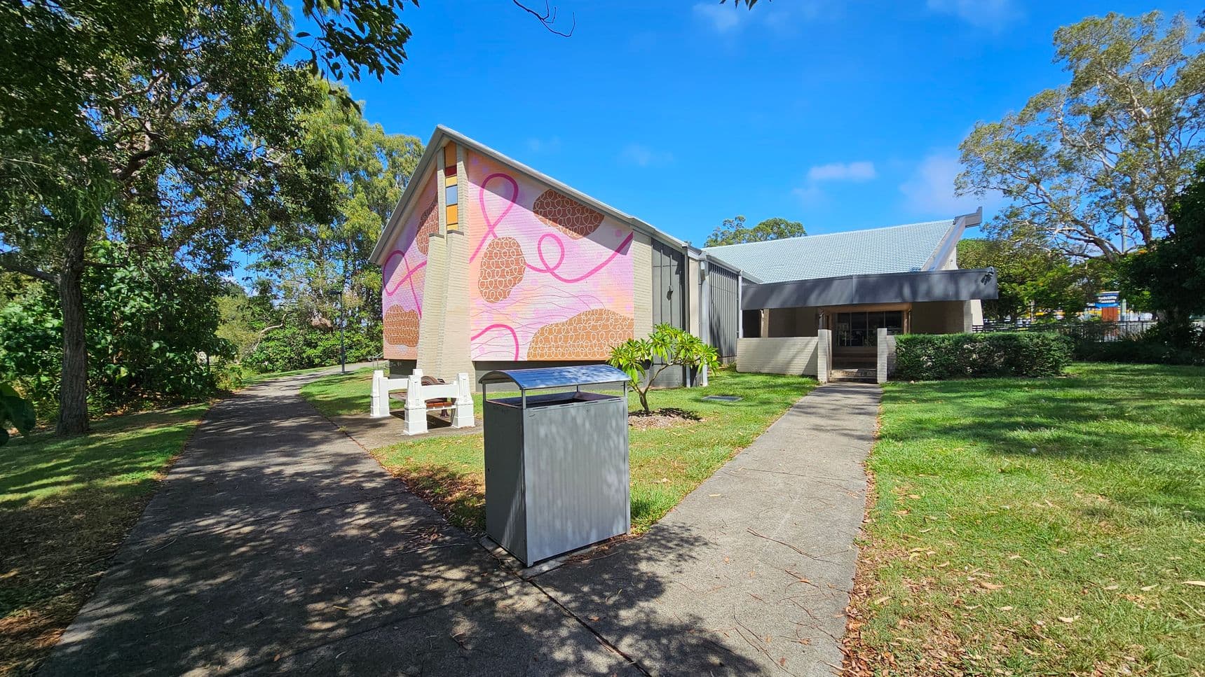 A building with a sloped roof and a colourful mural on the side wall featuring pink and brown abstract patterns. In the foreground, a paved path leads towards the building, with a metal bin on the right. On the left, there are trees providing shade over the path. The grass surrounds the path and building, with a few small bushes near the entrance. The sky is clear and blue.