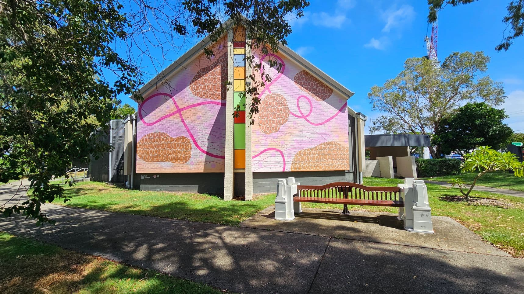 A building with a large, colourful mural on its side, featuring abstract patterns in pink, orange, and brown. In front of the building, there is a wooden bench on a concrete path. Trees with green foliage are on the left and right, providing shade. The grass is neatly trimmed, and a tall structure, possibly a tower, is visible in the background. The sky is clear and blue.