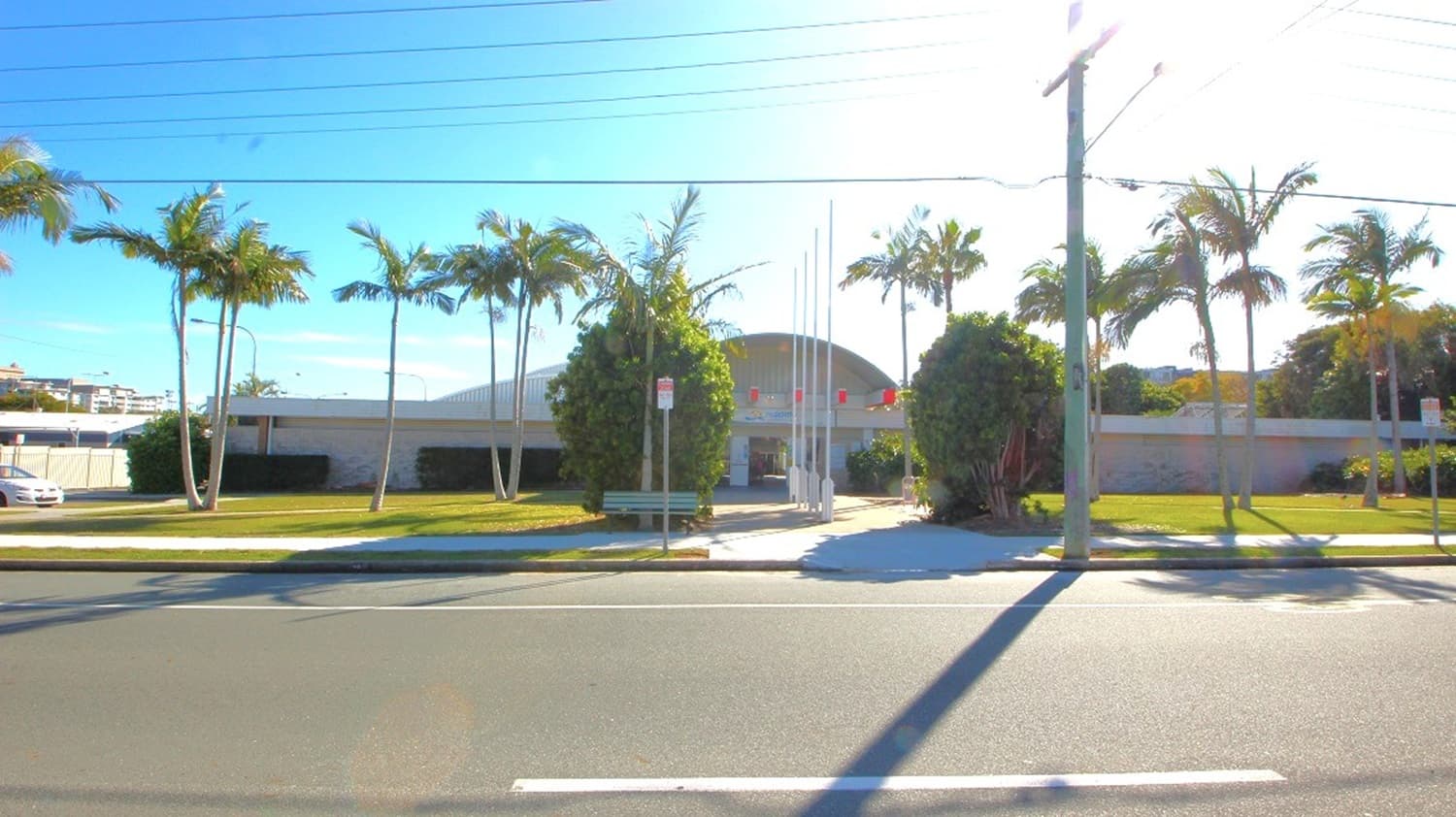 The Redcliffe War Memorial Swimming Pool is close to Sydney Street and is landscaped with rustling palm trees, one of which has a bench underneath it, shade-giving hedge plants, lush green grasses, and four white flagpoles lined up. The building entrance is symmetrically placed in the middle of the two separate front long walls made of bricks.