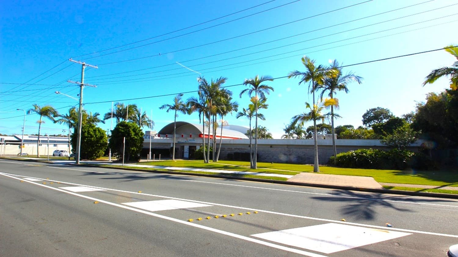 The road surface marking on Sydney Street, where the location is, indicates that the Redcliffe War Memorial Swimming Pool is close to the main road. The front of the building is surrounded by palm trees, hedges, and walkways.