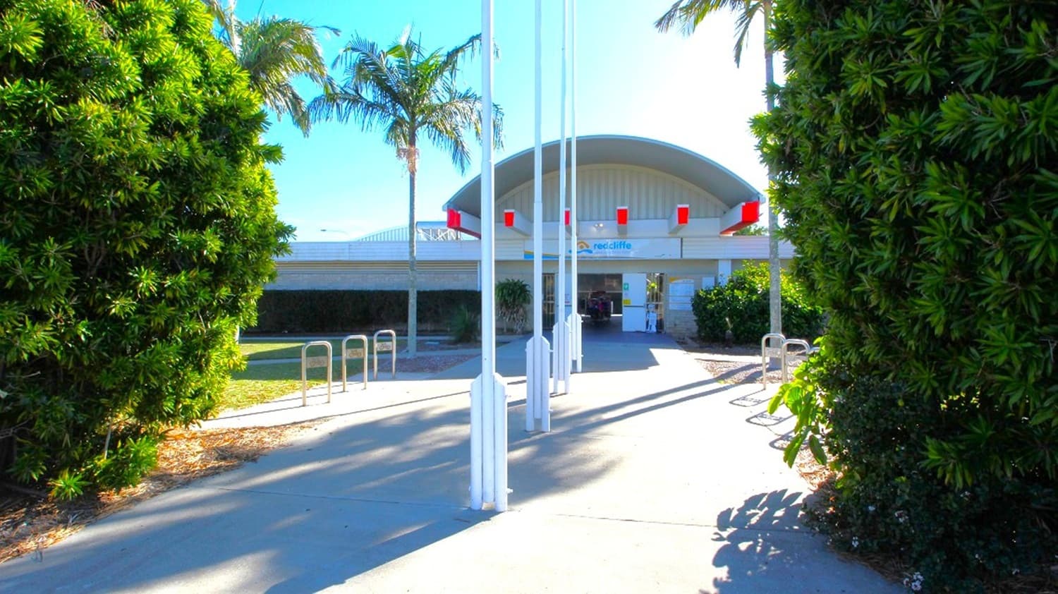 There are four white flagpoles lined up and three hoop barriers on either side of the path leading to the main entrance, and signage with the name "Redcliffe War Memorial Swimming Pool" is visible on the entrance's upper corner.