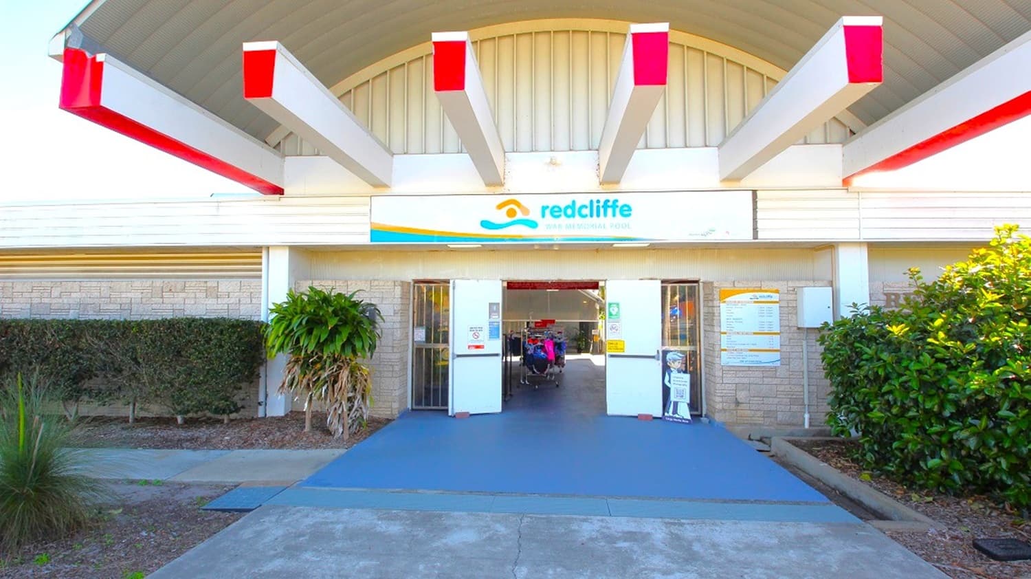 Entrance to a building with a sign reading "Redcliffe" above the door. The structure features a series of red and white beams extending from the roof. The entrance is flanked by plants on both sides, with a bush on the left and a hedge on the right. The ground is paved with a blue walkway leading to the door. The walls are light-coloured with a textured pattern.