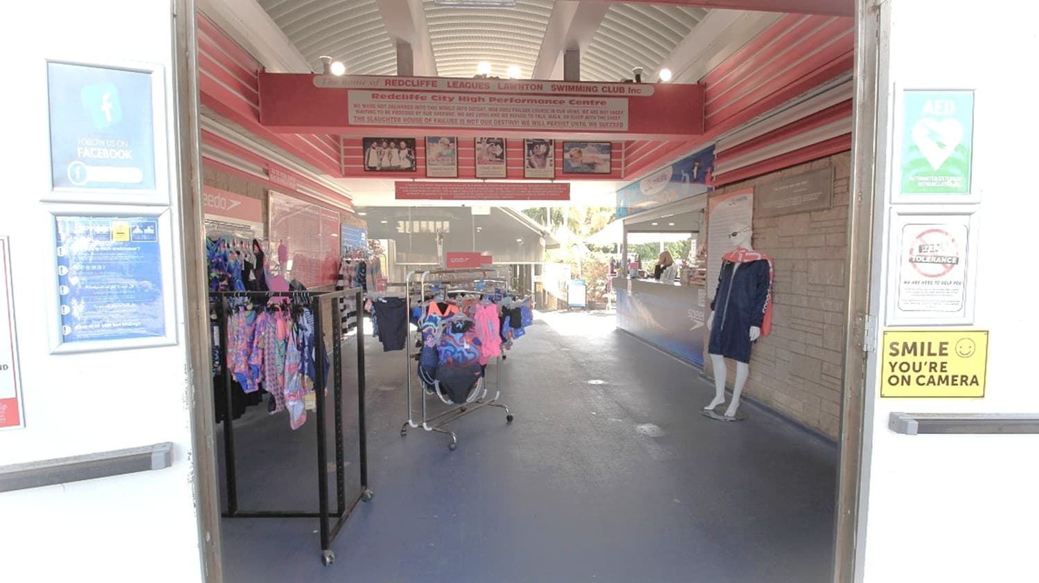 Entrance to a shop with clothing racks displaying colourful garments on the left. A mannequin dressed in a dark outfit stands on the right. Signs and notices are posted on the walls, including a "Smile, You're on Camera" sign on the right. The ceiling is arched with lights, and the floor is dark. In the background, more clothing displays and a counter are visible, with natural light coming from outside.