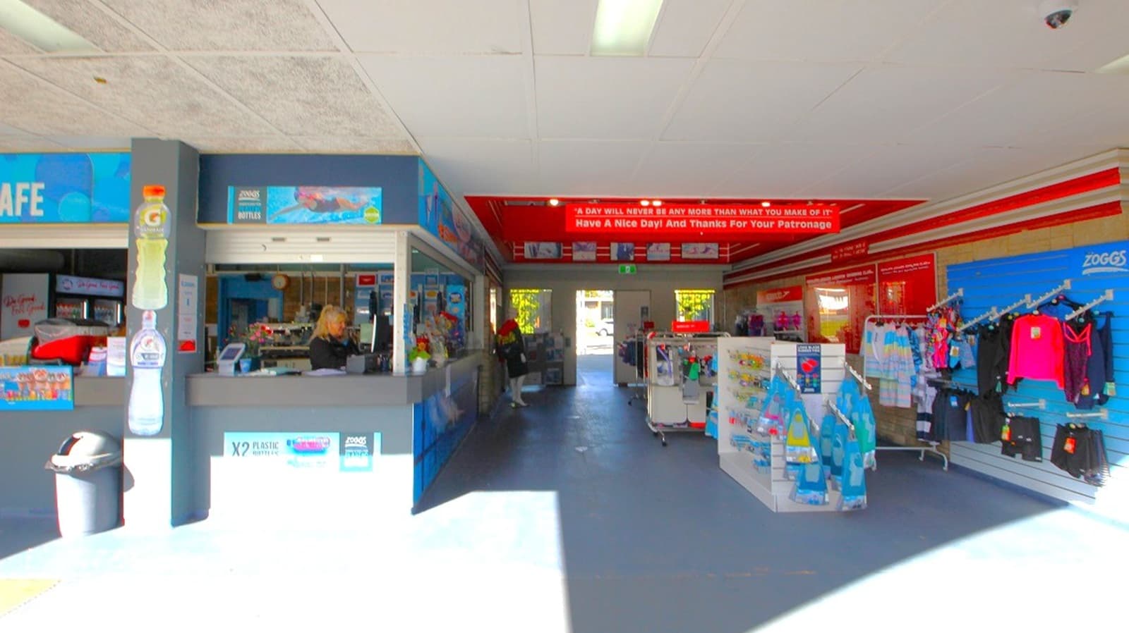 A shop interior with a blue and grey counter on the left, where a person is standing. The counter has signs and a bin nearby. In the centre, there is a walkway leading to a door with natural light coming through. On the right, shelves and racks display clothing and other items. The ceiling is white with fluorescent lighting, and the floor is a dark grey. A red sign with white text is visible above the walkway.