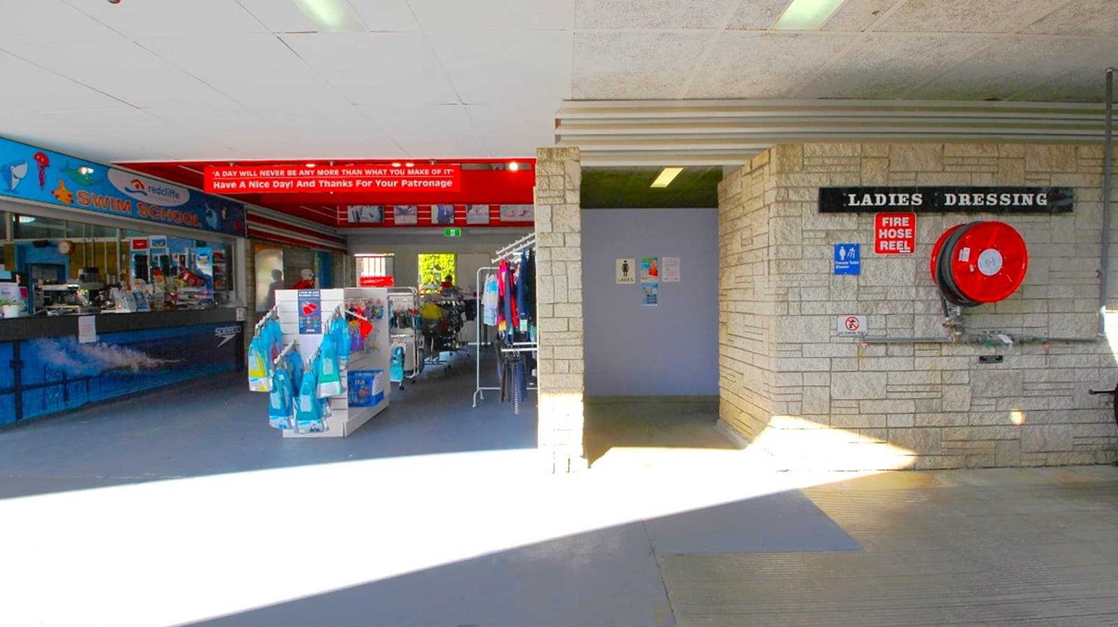 On the left, there is a shop with clothing racks and shelves displaying various items. A sign above reads, "Have a nice day! And thanks for your patronage." On the right, a brick wall features a sign for "Ladies Dressing" and a fire hose reel. The entrance to the dressing area is visible, with a small corridor leading inside. The floor is tiled, and the ceiling has fluorescent lights.