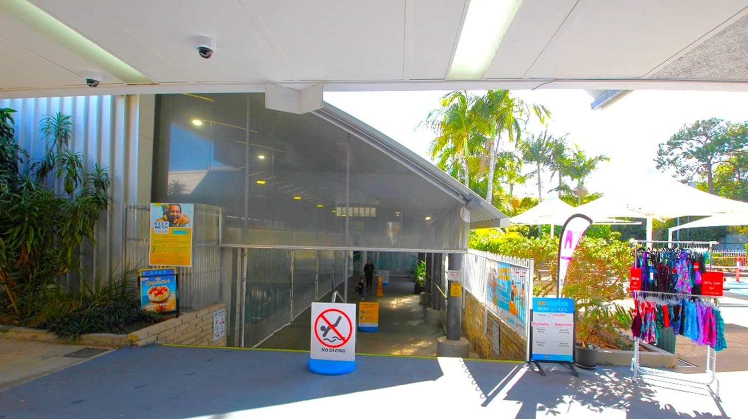 Entrance to a building with a sloped roof and glass facade in the centre. A sign with a no-smoking symbol is in front. On the left, there are posters and plants. On the right, a rack displays colourful clothing, and there are white umbrellas and palm trees in the background. The ceiling is white with lights, and the floor is grey.