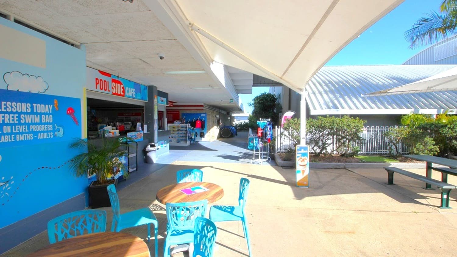Outdoor seating area with round wooden tables and blue chairs in the foreground. To the left, a blue wall with signage advertising swimming lessons and a poolside café. In the middle, a shaded walkway leads to a shop with various items on display. The background features a large, curved glass structure and some greenery. The ceiling is a white canopy providing shade.