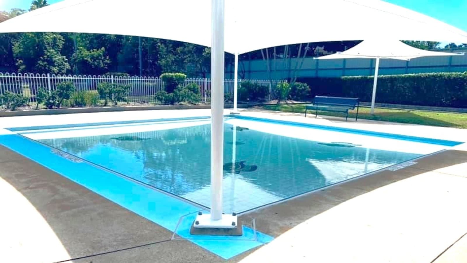 A small, shallow swimming pool with clear water is in the centre, surrounded by a light-coloured concrete deck. A large white shade sail is suspended above, supported by a central pole. In the background, there is a white fence with greenery and trees behind it. To the right, a bench and another shade structure are visible on a grassy area.