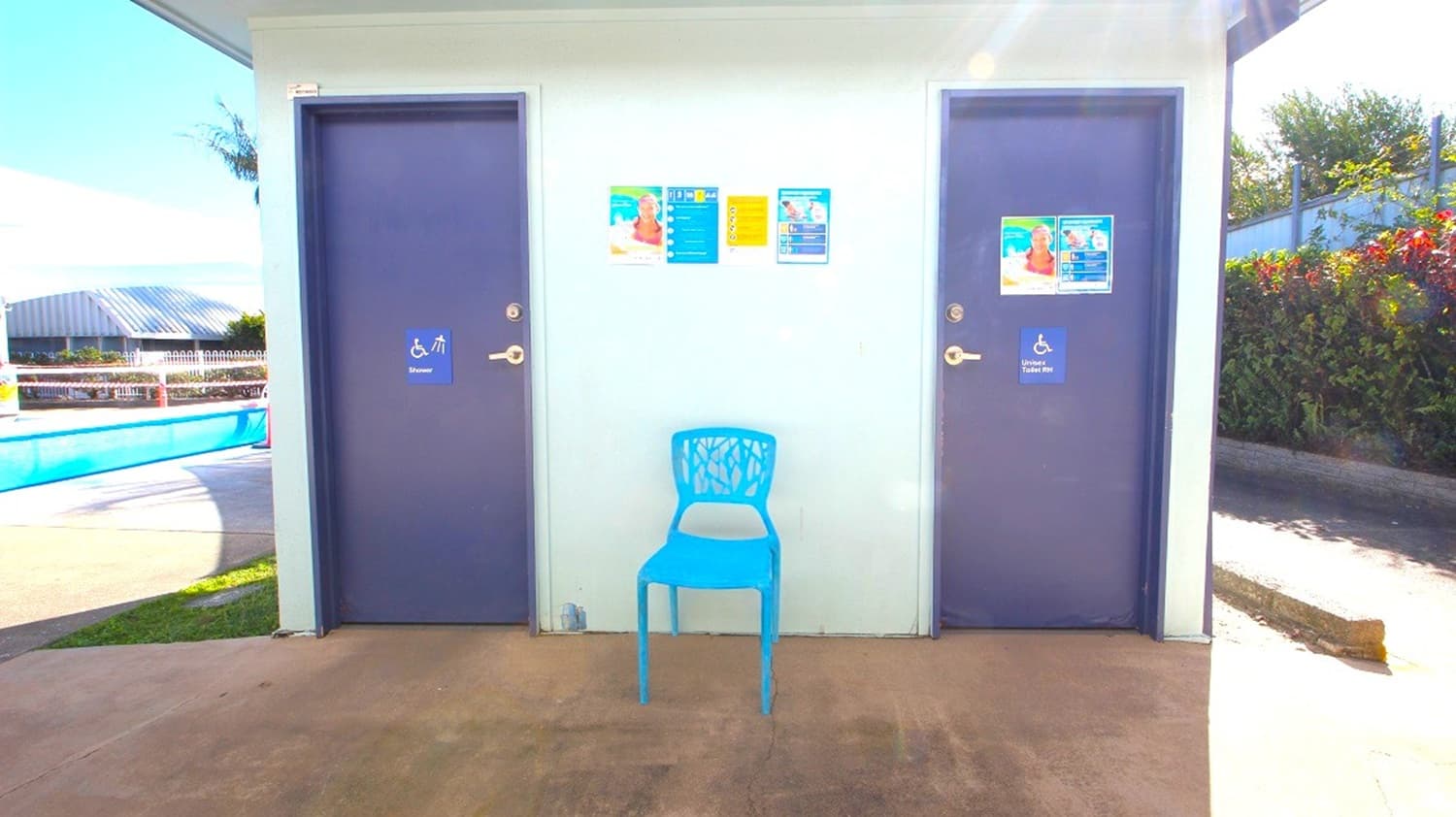 Two dark blue doors with disability signs are on either side of a light-coloured wall. In the centre, a blue chair is placed against the wall, which has several colourful posters. To the left, part of a swimming pool and a fence are visible. On the right, there is a hedge with red flowers. The ground is concrete, and the ceiling is not visible.