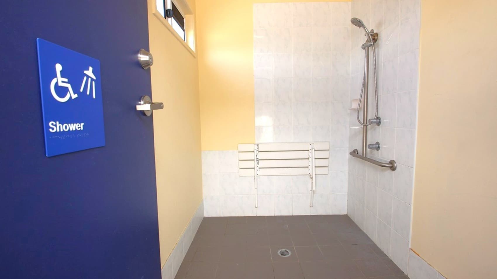 Accessible shower room with a dark blue door on the left, featuring a sign with a wheelchair symbol and the word "Shower". Inside, the shower area has a grey floor with a drain, a white wall with a foldable seat, and a showerhead with an adjustable hose. There are grab bars on the wall for support. A small window is visible above the door, allowing natural light into the space.