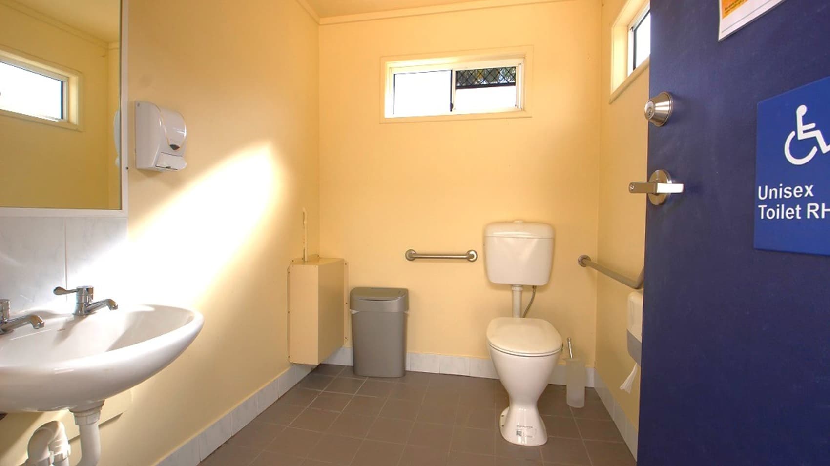 A bathroom with beige walls and a brown floor. On the left, there is a white sink with dual taps and a mirror above it. A small window is above the sink. In the centre, a toilet with a white cistern is flanked by metal grab bars. A small bin and a wall-mounted dispenser are nearby. On the right, a blue door with a sign indicating a unisex accessible toilet. Another small window is above the toilet. The ceiling is plain and light-coloured.