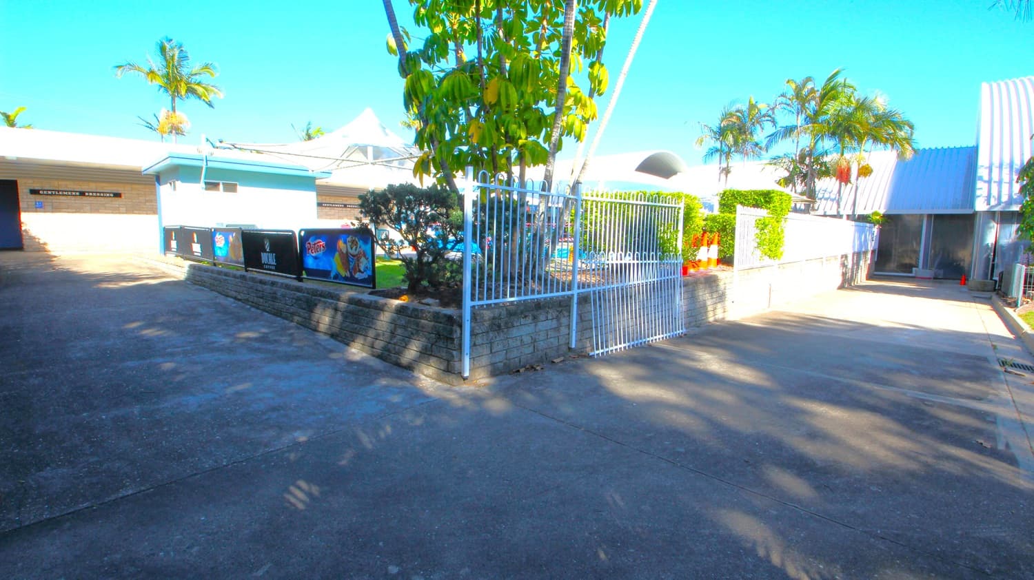 A paved area with a white metal gate in the centre, leading to a building with a sign that reads "Gentlemen's Dressing" on the left. The building has a light blue and white exterior. To the right, there is a pathway with orange traffic cones and a white fence. Several palm trees and a large leafy tree are visible, providing some shade. The ground is concrete, and the sky is clear.