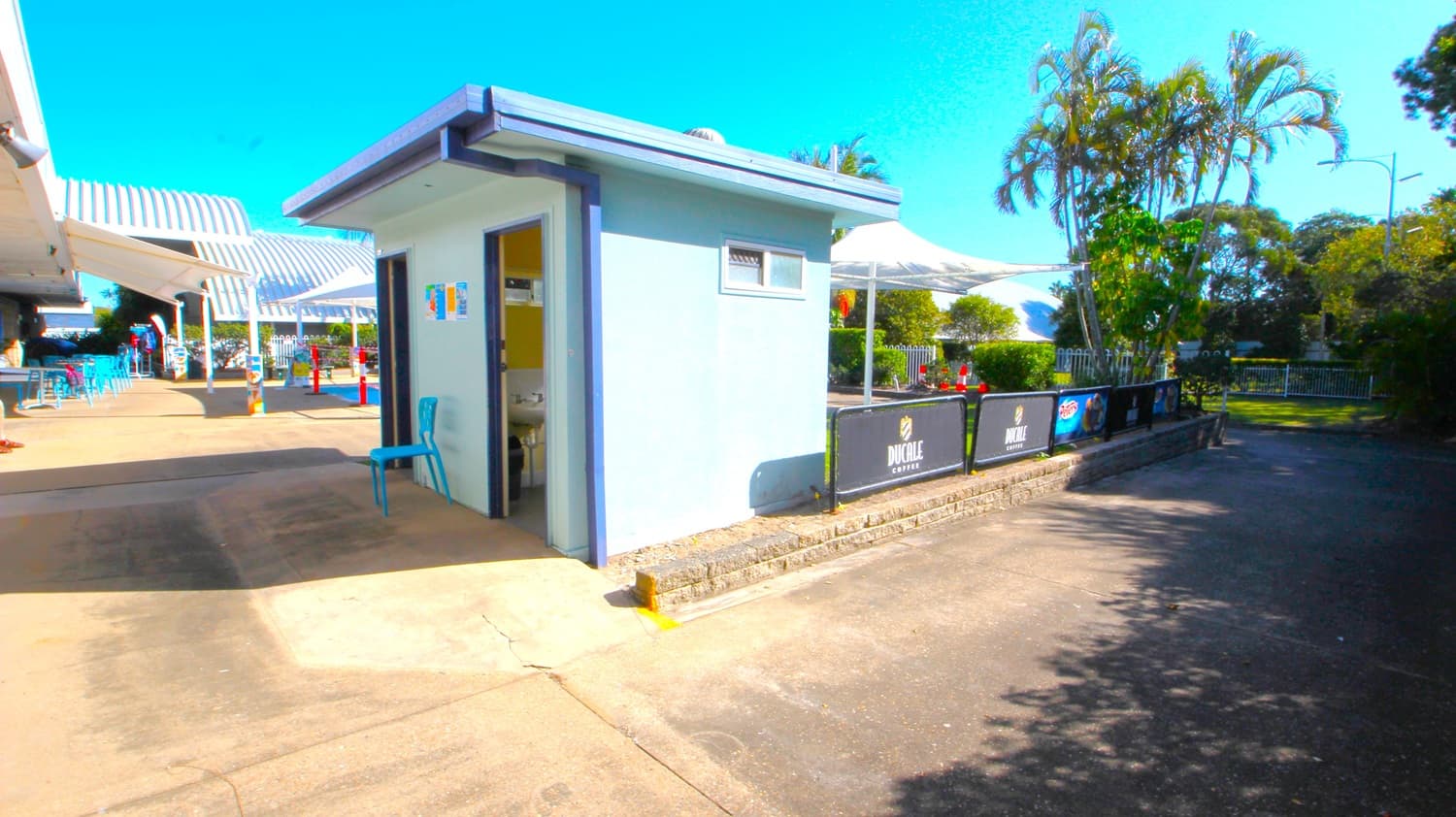 A small, light blue building with a flat roof is in the centre, featuring an open door and a small window. To the left, there are outdoor seating areas with blue chairs and tables under striped awnings. On the right, a path leads past a fence with advertisements, and there are tall palm trees and greenery. The ground is paved, and the area is well-lit by sunlight.
