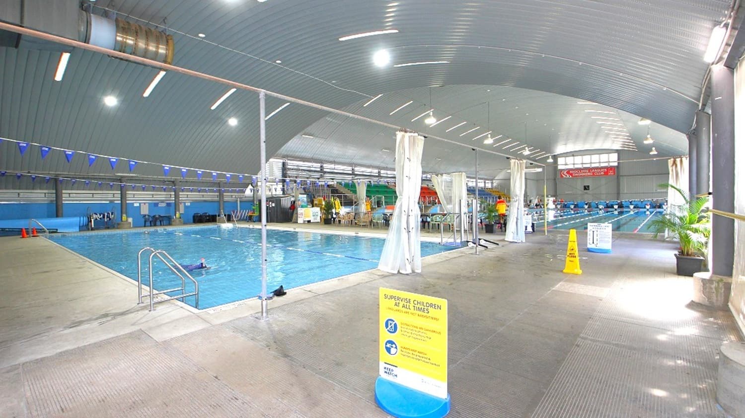 Indoor swimming pool area with a large pool on the left, featuring lane dividers and a metal handrail. The right side has a walkway with safety signs and potted plants. The ceiling is curved with multiple lights, and the background shows seating and more pool lanes. The floor is tiled, and there are white curtains hanging from poles along the walkway.