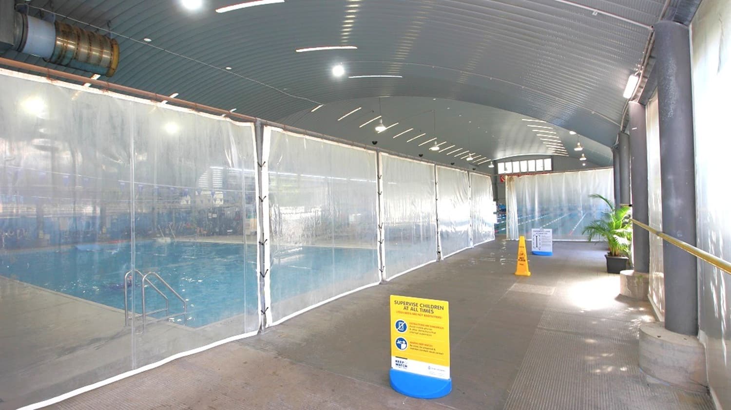 A covered walkway with a corrugated metal ceiling and a textured floor. On the left, a transparent barrier reveals a swimming pool with visible water and pool steps. In the centre, a yellow sign with safety instructions is placed on the floor. On the right, a potted plant is near the wall, and a caution sign is visible further down the walkway. The area is well-lit with overhead lights.