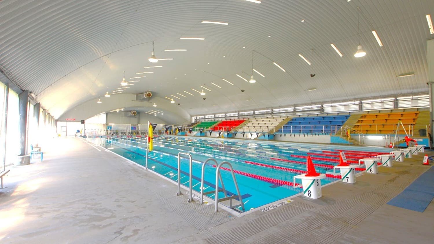 Indoor swimming pool with multiple lanes, each marked by red lane dividers. On the right, there are tiered seating areas with blue, red, and yellow seats. The left side features large windows allowing natural light to enter. The ceiling is curved with evenly spaced lights. The pool deck is wide, with metal ladders for pool access and orange cones near the starting blocks. The floor is tiled, and the overall space is spacious and well-lit.